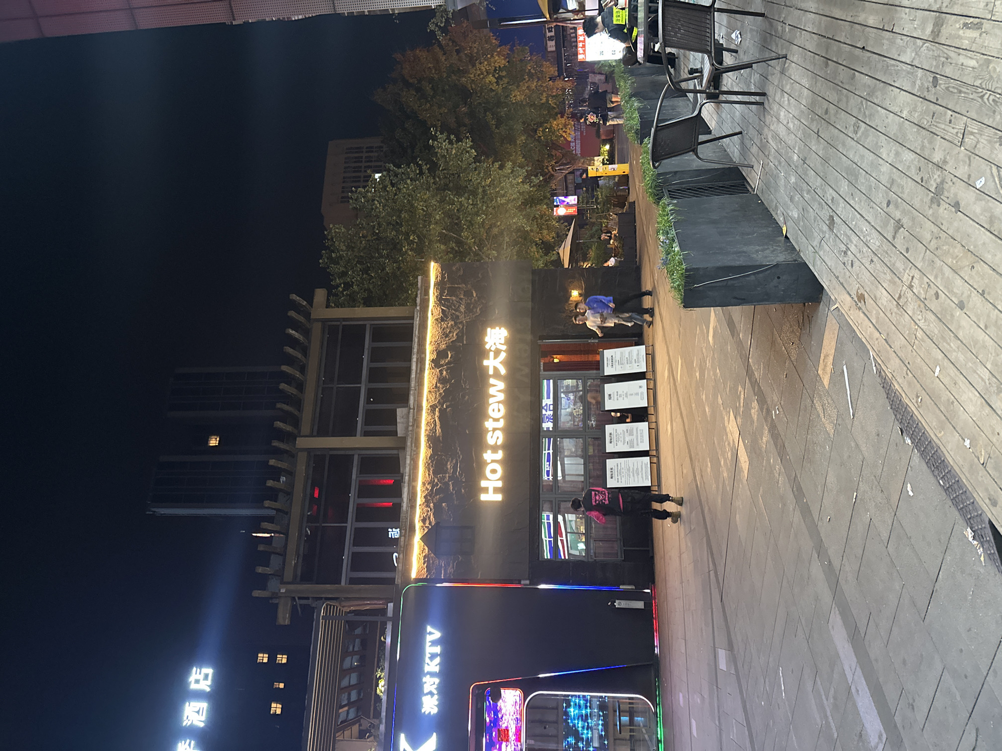 The image depicts a night scene in the Chongqing High-tech Zone of China. A building prominently features the words "Hot Stew 大火". A KTV establishment is also visible. People are walking on the sidewalk. Outdoor seating with chairs and planters is present. Buildings and trees are in the background.