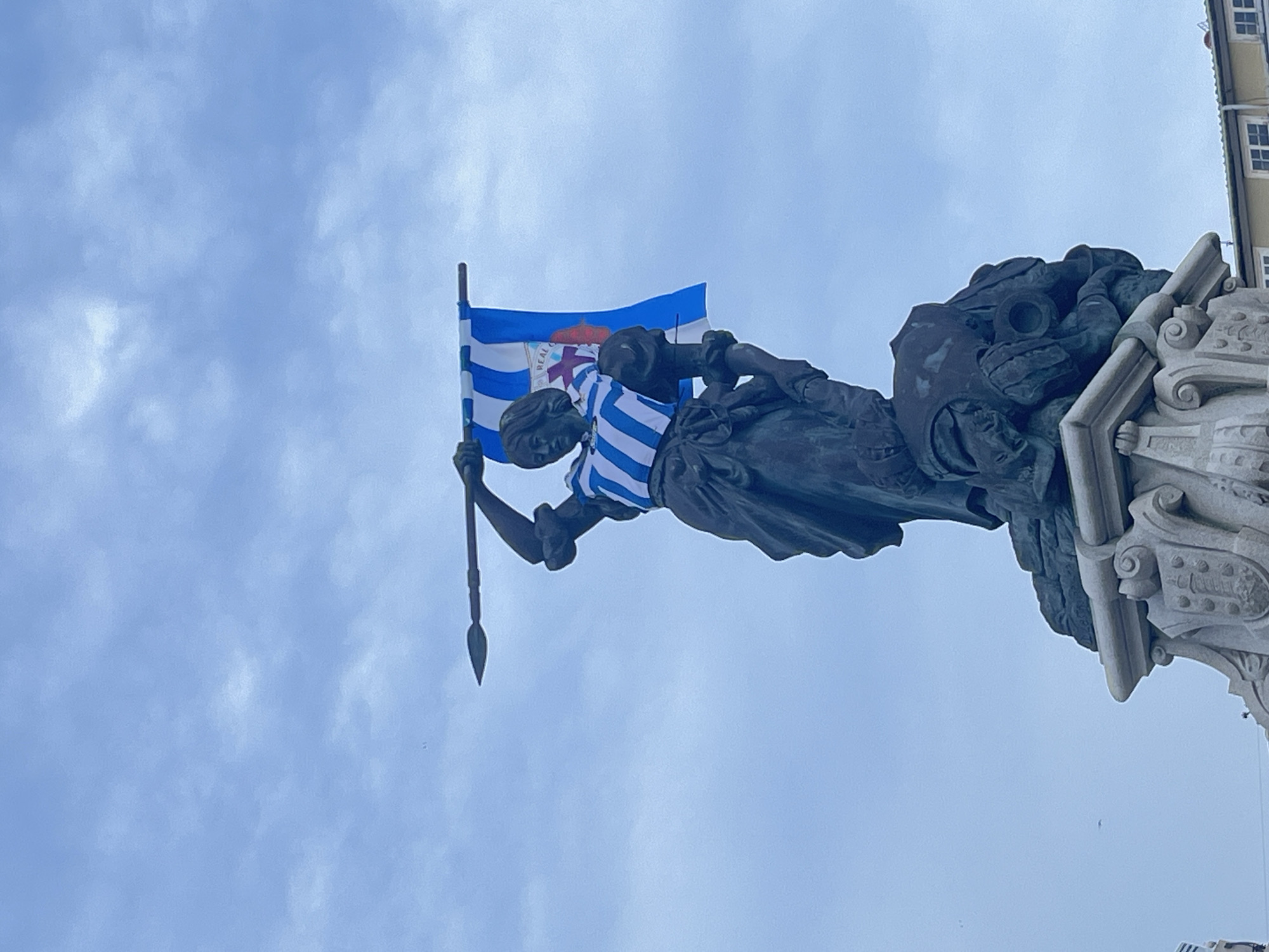 A low-angle shot captures a bronze statue of a woman holding a spear, located in A Coruña, Spain. The statue is adorned with a blue and white striped jersey around its upper torso. A flag with blue and white stripes, possibly of a local sports team, is held aloft by the statue. The background consists of a cloudy sky and a corner of a building with windows visible in the upper right of the image. The statue is mounted on an ornate stone base.