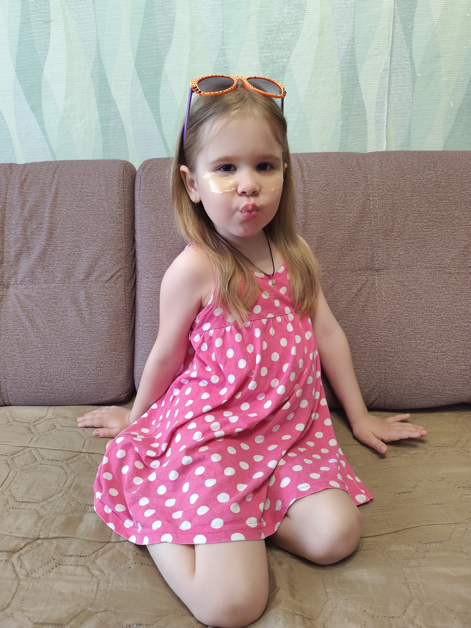 A young girl sits on a brown couch. She wears a pink dress with white polka dots and has orange sunglasses on her head. There are small, translucent patches on her cheeks. The background consists of a green and white patterned wall. The scene likely takes place indoors in Фоки, Russia, based on the location information provided.
