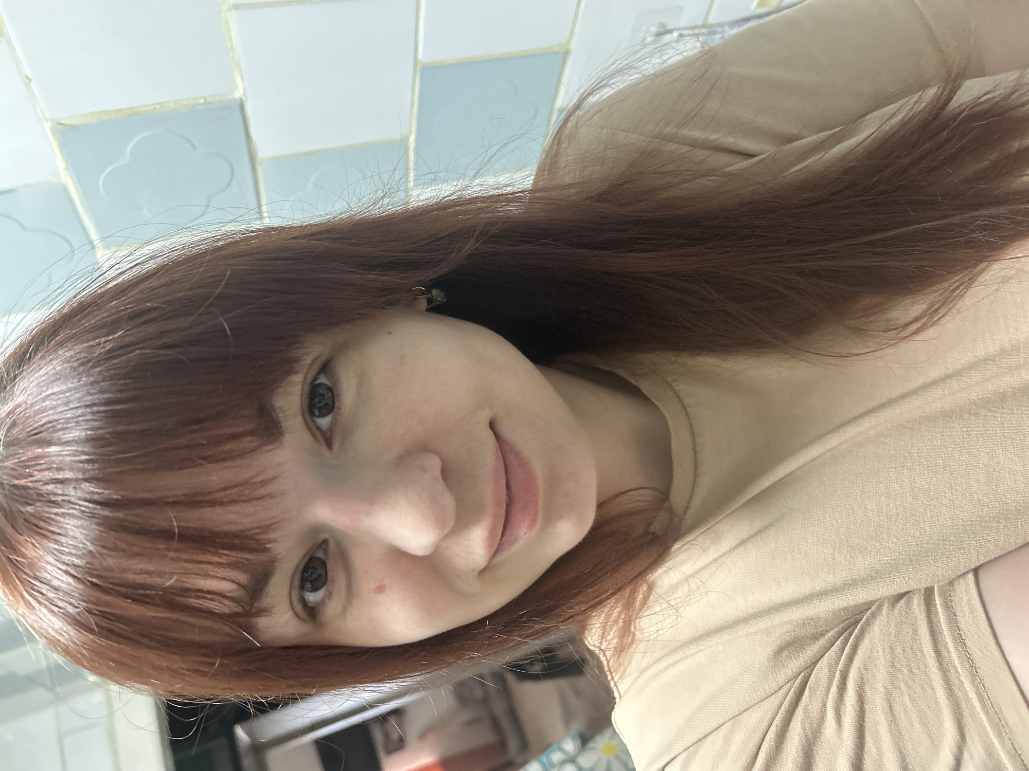 A selfie of a light-skinned woman with reddish-brown hair and bangs. She is wearing a tan shirt. The background includes light blue and white tiles. The location cannot be determined as Фоки, Russia, with the available information.