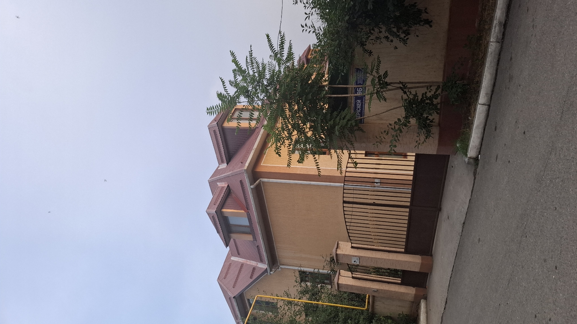 This is a vertical, full-shot photograph of a house and its surroundings on a street. The house is a two-story building with a reddish-brown, tiled roof and beige-colored walls. It features multiple windows, some of which are dormer windows in the roof. A brown metal gate is visible in front of the house, along with a concrete fence and a paved driveway. Trees and foliage surround the property, with a large tree partially obscuring the house. A bright yellow gas pipe runs along the side of the house. The sky is overcast and appears to be a light blue or grey, suggesting it is daytime, possibly early morning or late afternoon, with overcast weather. There are a few small, blurry objects in the sky that might be birds or kites. A street sign with blue lettering and white text is mounted on the wall of the house, partially obscured by the tree. The text on the sign appears to read "ЕРІСКЕЙ 95". The image is oriented vertically, with the sky at the top and the street at the bottom. The overall scene depicts a residential area.