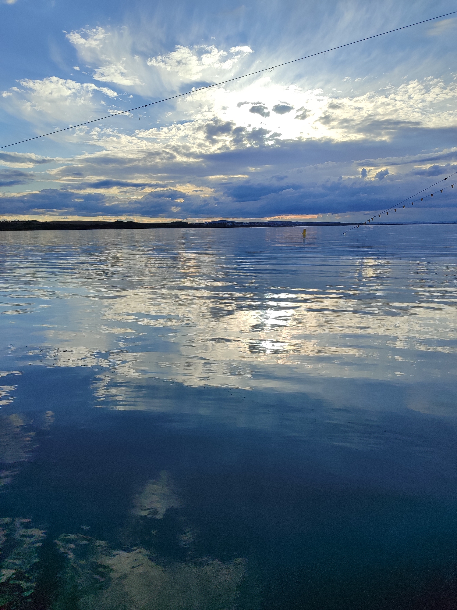 The image depicts a body of water under a cloudy sky. The water's surface reflects the clouds and the bright, presumably setting, sun. In the distance, there appears to be land, possibly a shoreline. A thin line runs across the upper portion of the image, likely a wire or rope. Further to the right, a strand of small, triangular flags can be seen. A single yellow buoy floats on the water. The location could be a body of water in or near Burgas, Bulgaria. Details such as landmarks are not visible, but the general impression is of a coastal or lakeside area.