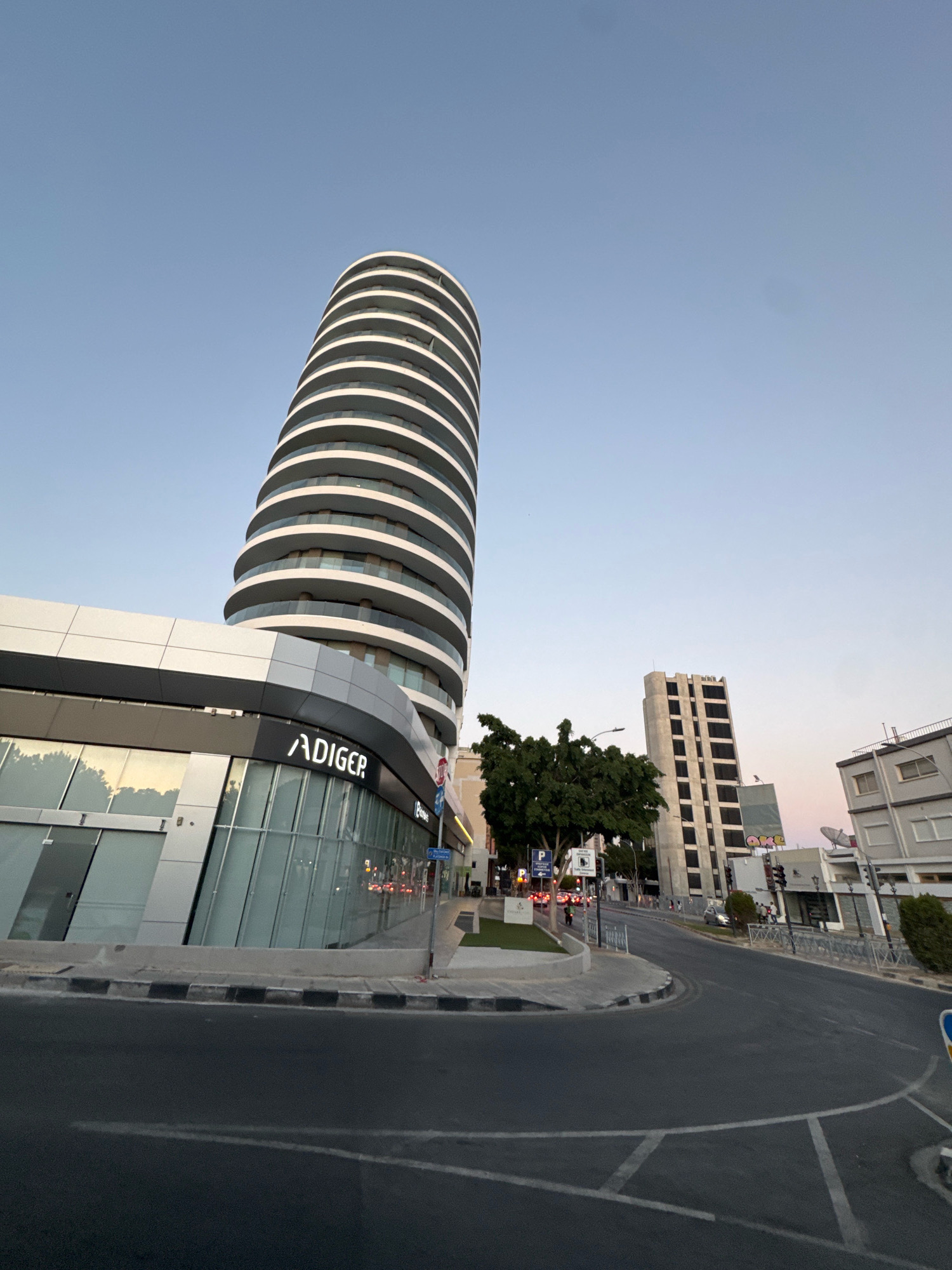 A wide-angle, low-angle shot captures a modern, curved skyscraper with horizontal white balconies against a clear blue sky. In the foreground, a contemporary building with a glass facade and the prominent "ADIGER" logo is visible. The scene depicts a street in Limassol, Cyprus, with a road curving to the right, bordered by sidewalks and streetlights. Other buildings, including a taller, rectangular structure with dark windows, are present in the background. The lighting suggests it is either early morning or late afternoon. There are no prominent human figures in the image.