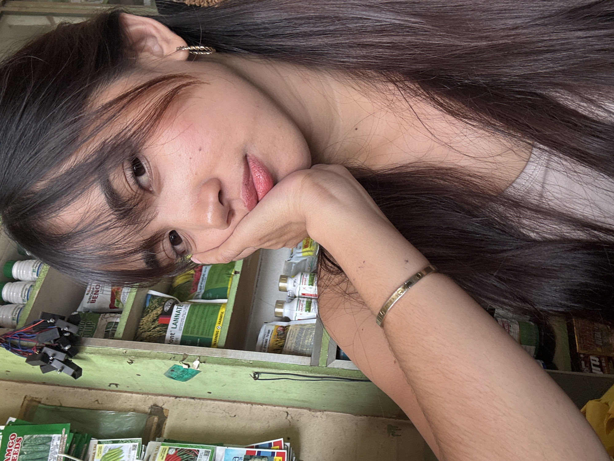 The image shows a young woman resting her right hand on her cheek, her left arm is bent and her hand is near her chest. She has long dark hair with bangs and is wearing a light-colored top and a gold-toned bracelet. She is looking towards the camera with a slight smile. The background is cluttered with shelves filled with various items, including bags of seeds and bottles, suggesting a retail environment. The overall impression is that of a casual selfie taken in a small shop. It is not possible to identify the exact location in Tagum City from the image alone.
