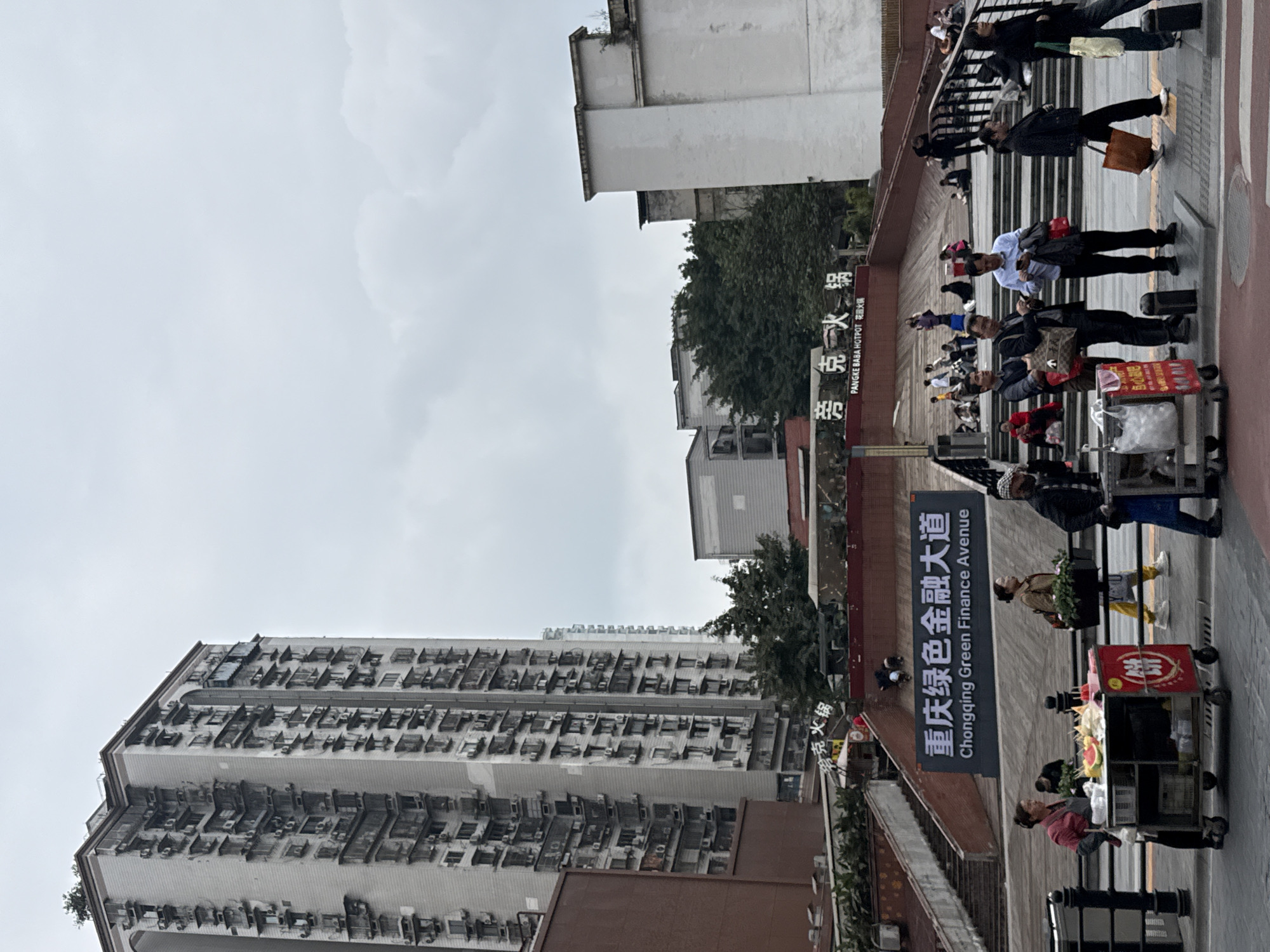Here is a description of the image:
The photograph depicts a street scene in Yuzhong District, Chongqing, China. A tall, high-rise apartment building dominates the left side of the image. Many individual air conditioning units are installed on the exterior of the building. Overhead sky is overcast. A pedestrian walkway runs along the right side of the image, with people walking and wheeling carts with goods. A sign indicates "Chongqing Green Finance Avenue". Text in Chinese is also on the sign. Additional buildings and trees are visible in the background. "PANGKE BABA HOTPOT" text can also be seen, and is associated with a restaurant.