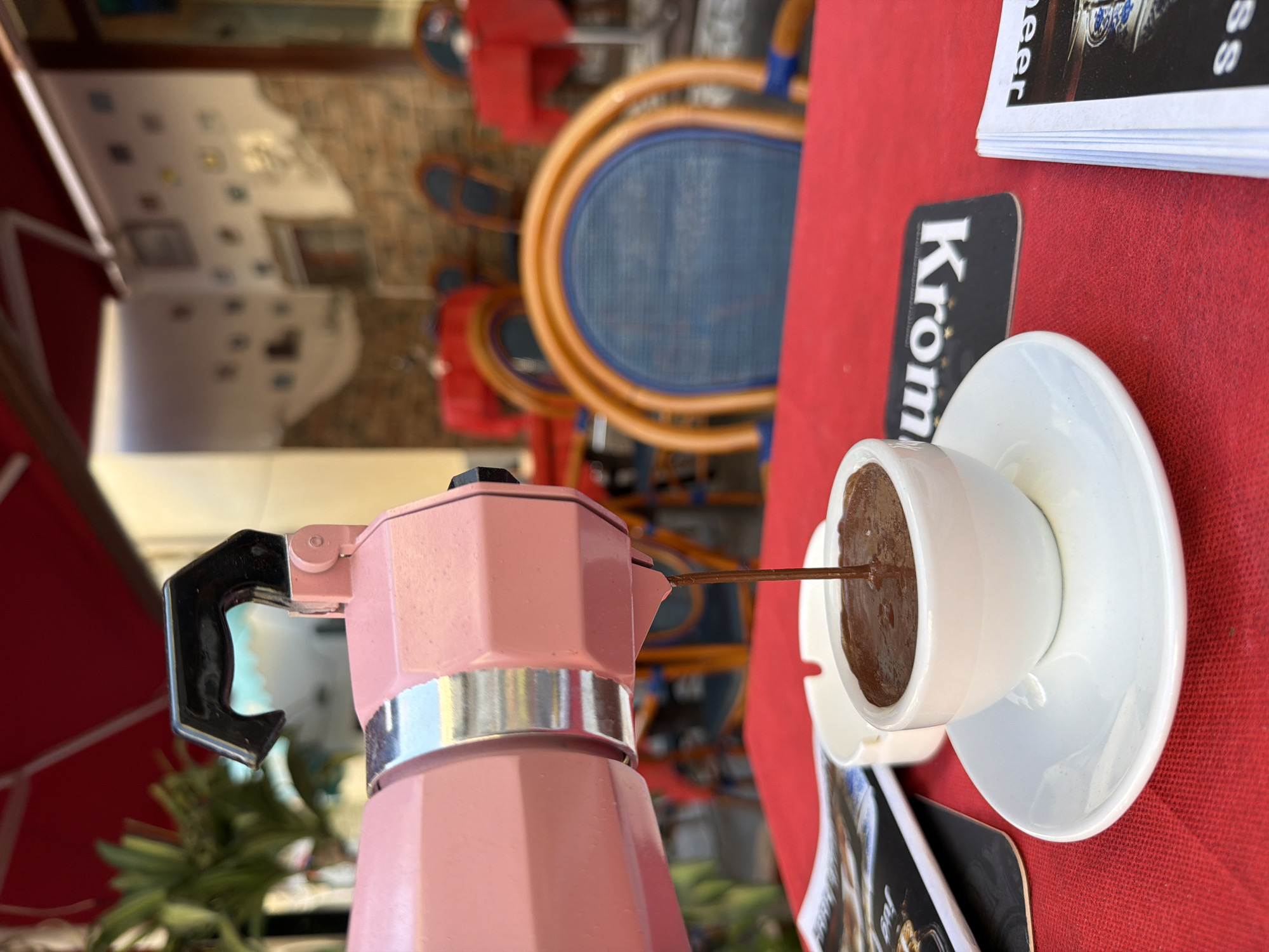 A Moka pot pours coffee into a cup. The pot is pink with a black handle, while the cup and saucer are white. A dark grey beer coaster and a stack of beer promotional cards sit on a red tablecloth. In the background are several blue and rattan dining chairs with red seats. The scene is outdoors in Ischia, Italy.
