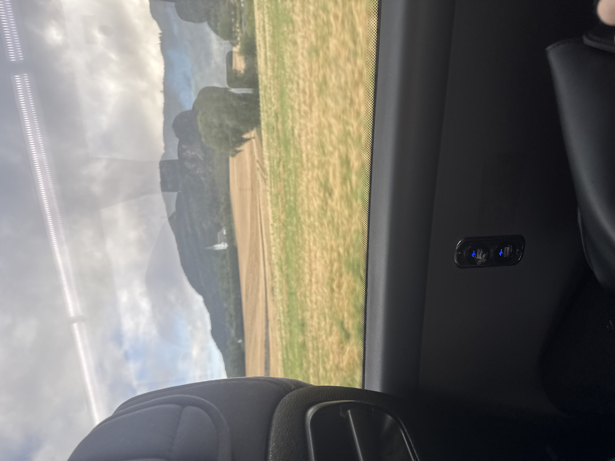 An interior shot from a vehicle, likely a train, shows a view through the window of a rural landscape. In the foreground, the back of a gray fabric headrest is visible on the left. Below the window, a dark gray plastic panel houses two USB ports, both illuminated with a blue light. The window reveals a sunlit field of yellowing grass or crops, bordered by a green field. In the distance, rolling hills and mountains are silhouetted against a cloudy sky. A white steeple, possibly of a church, is visible amongst the trees in the middle ground. The location is identified as Skatval, Norway.
