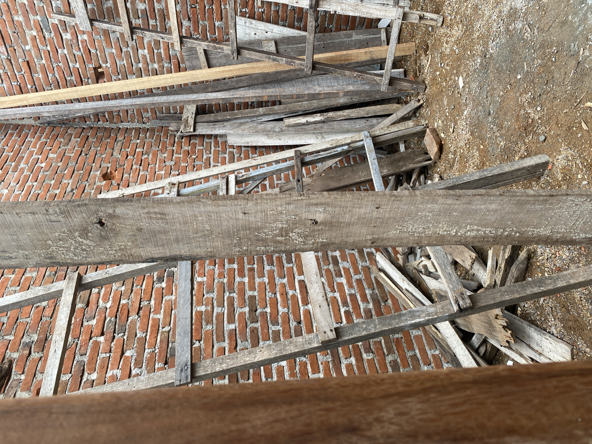 A close-up, eye-level shot focuses on a brick wall and wooden scaffolding at a construction site. The brick wall, constructed from reddish-orange bricks with white mortar, dominates the background. Leaning against the wall are various pieces of unfinished wooden lumber, some appearing weathered and splintered. A rudimentary wooden ladder structure, made of rough-hewn planks, is positioned in the foreground and partially obscures the view of the wall. The ground beneath is covered with dirt, gravel, and scattered pieces of wood debris, suggesting an active or recently worked construction area. The scene is dimly lit, casting subtle shadows that add depth and texture. No individuals are visible in the frame.