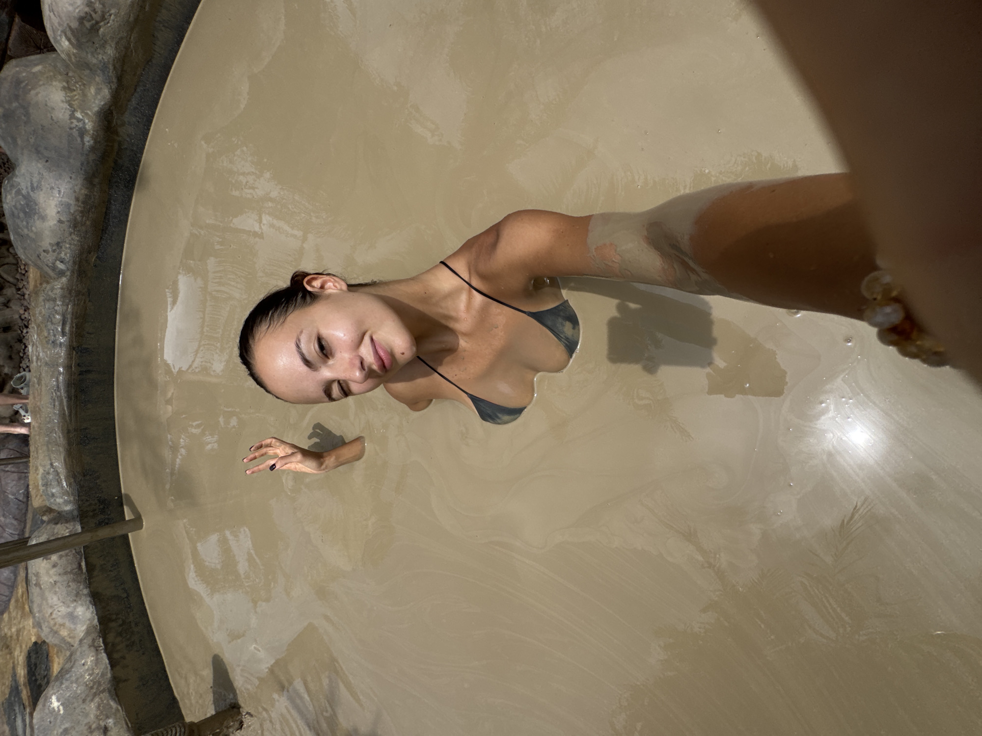 A woman is pictured from an overhead perspective, partially submerged in a beige, mud-like substance within a circular pool. She has dark hair pulled back and is wearing a dark-colored string bikini. She is extending one arm towards the camera, the other is submerged up to her wrist, near her shoulder, and she is smiling slightly. The pool is edged with a dark, irregular stone border. A portion of what appears to be a wooden or metal railing is visible in the bottom left corner. The scene is likely from the mud spa at บ้านคลองเคียน, Thailand, based on the presence of the mud and the woman bathing.