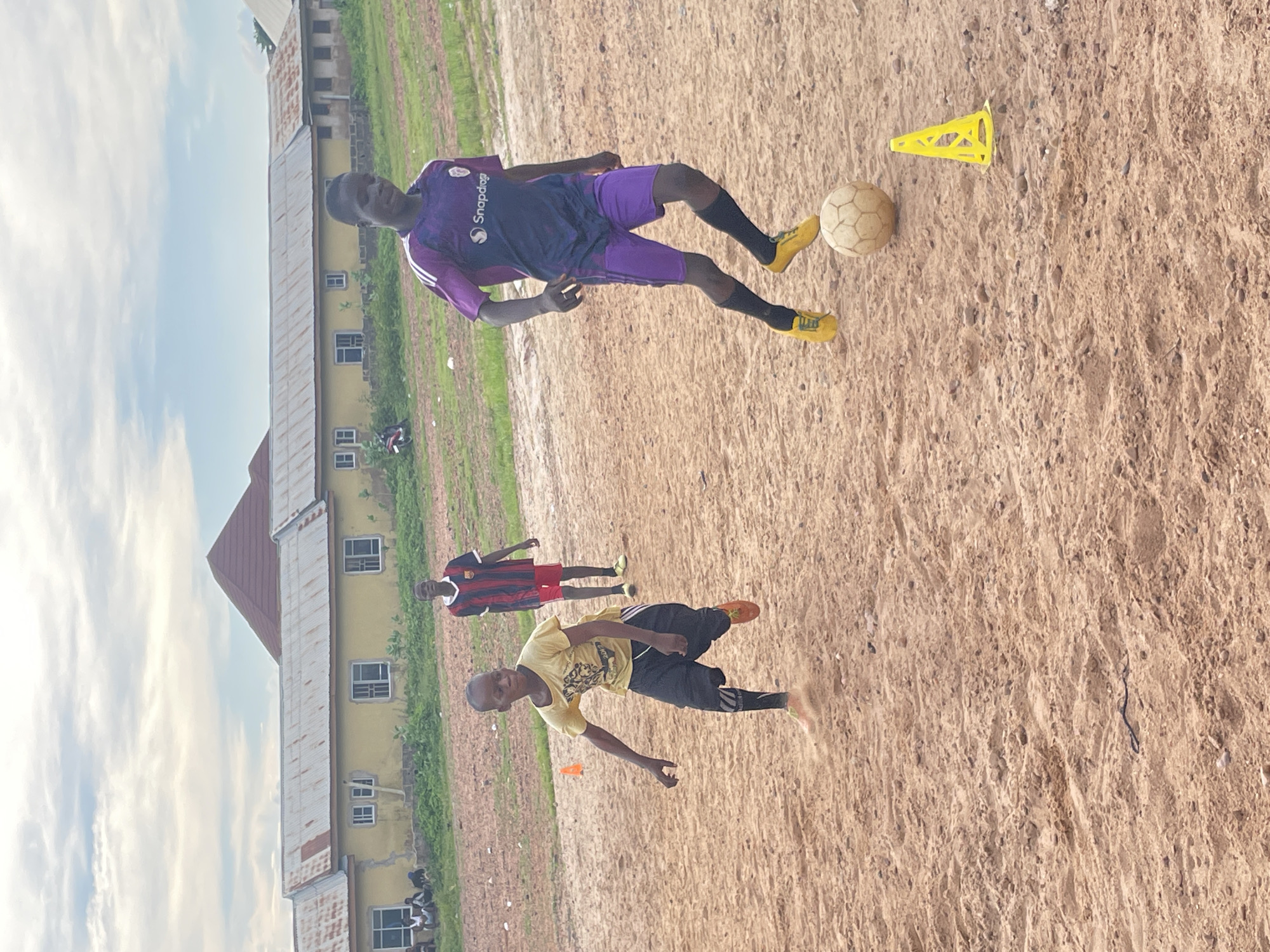 The image depicts an outdoor scene in Lokoja, Nigeria, likely a sports field with a dirt or sand surface. Three young males are visible. In the foreground, a boy in a yellow t-shirt and black shorts is running, kicking up dust. To his right and slightly behind him, another boy in a purple and dark blue soccer jersey and purple shorts dribbles a soccer ball. Further in the background, a third boy wearing a red and black striped soccer jersey and red shorts stands observing. In the background, a long, low building with a corrugated metal roof and yellow walls is visible. The sky is overcast with some clouds. A yellow traffic cone is placed on the ground near the boy with the ball.