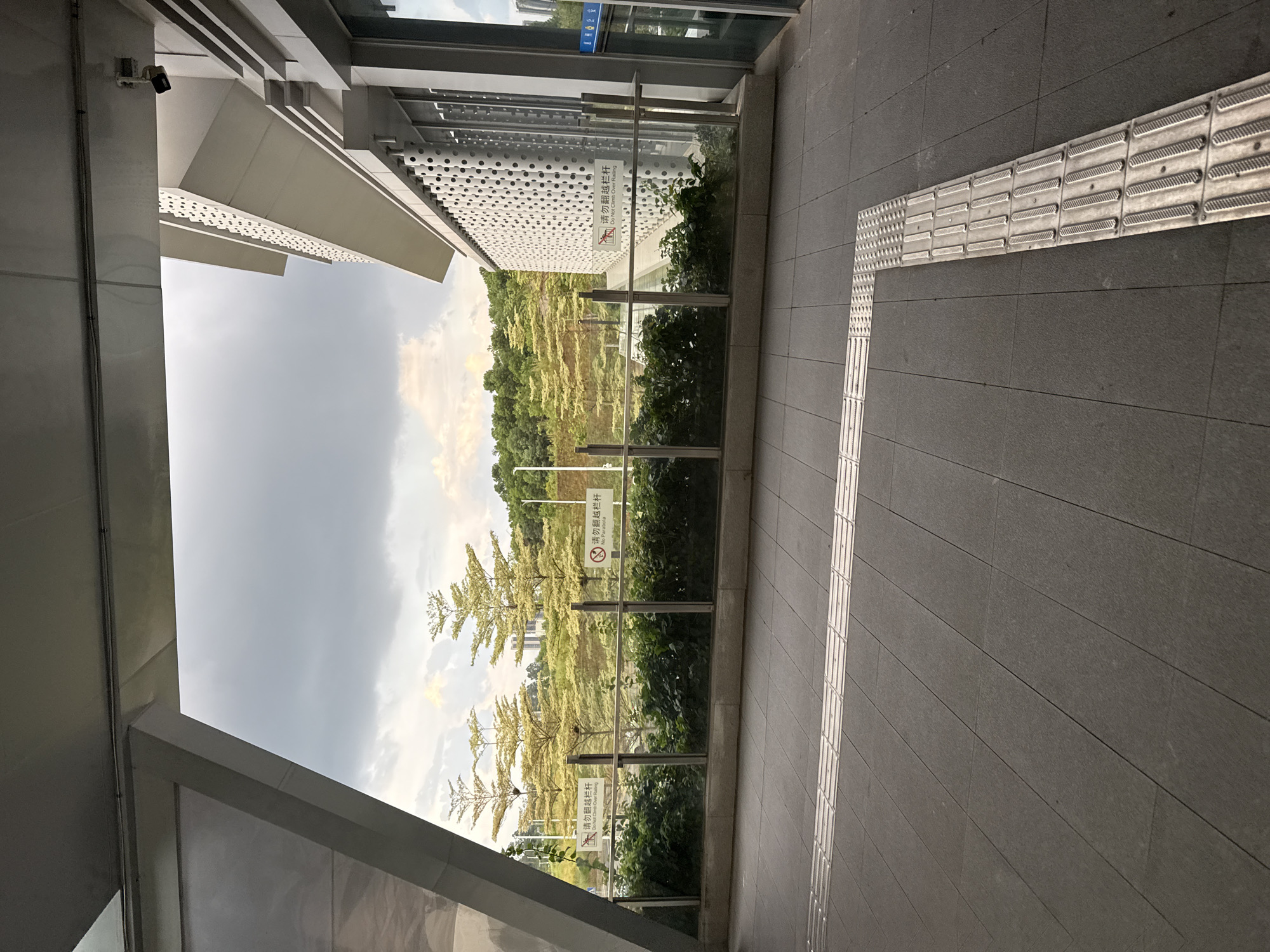 The image depicts an outdoor walkway with a tiled floor and tactile paving for the visually impaired. In the background, there is a lush green hillside dotted with trees and buildings. The scene appears to be from a public space, possibly a park or a transportation hub, given the modern architecture and the presence of warning signs. There are no people visible in the image. The sky is overcast, suggesting a cloudy day. The location is identified as Huizhou, China.