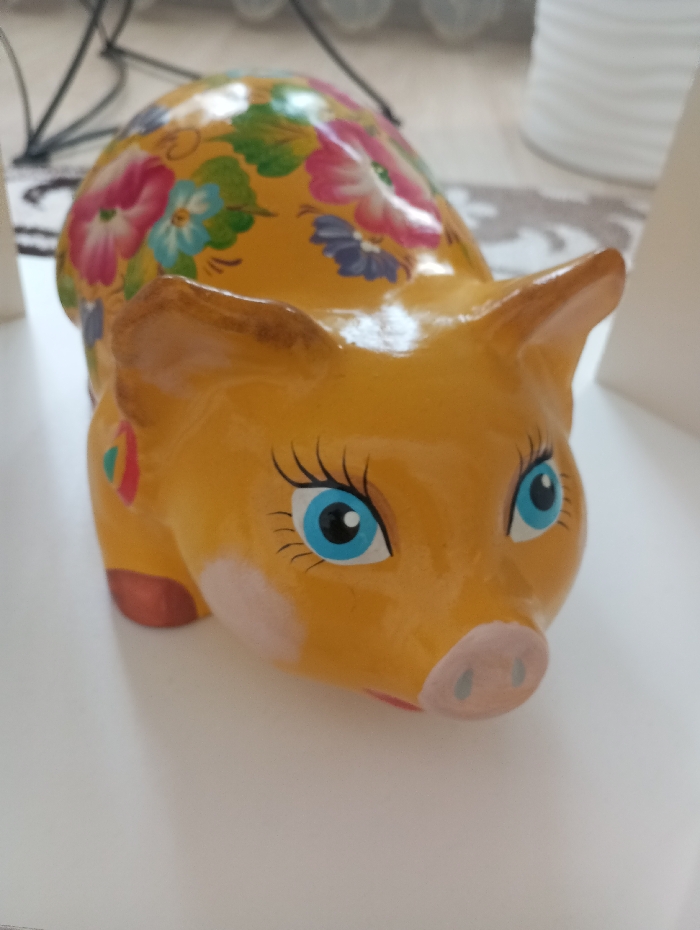 A yellow-orange, pig-shaped decorative object, possibly a piggy bank, rests on a flat, white surface. The object features painted blue eyes with black pupils and prominent black eyelashes, a pink snout with two dark nostrils, and light pink blush marks on its cheeks. Its ears and visible hooves are tipped with a light brown color. The main body is adorned with a floral pattern consisting of pink, blue, and purple blossoms with green leaves. A small, multi-colored (red, yellow, green) abstract mark is present on the object's left flank. The background is out of focus, showing indistinct dark linear elements and blurred white and patterned surfaces. The scene is situated in the city of Яромаска, Russia.