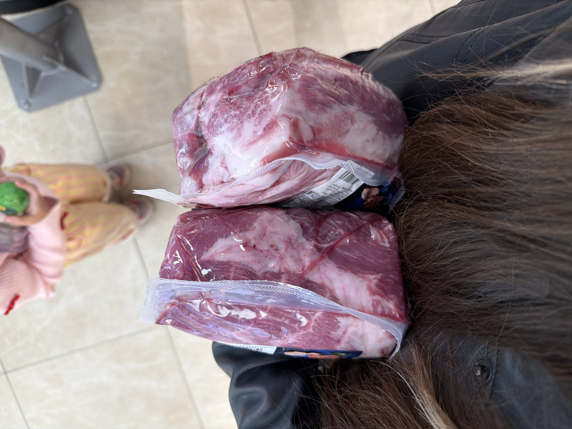 Here's a description of the image:
The image shows two packages of fresh meat, likely pork due to the color and marbling, stacked on top of each other. The meat is wrapped in clear plastic packaging. The packages are being held against the shoulder of a person wearing a dark leather jacket. Strands of the person's brown hair are also visible. In the background, there is a glimpse of a tiled floor, what appears to be the base of a shopping cart, and the legs of a person wearing what looks to be a pink and yellow outfit.
The image does not contain enough information to place the location in Moscow, Russia, or any city.