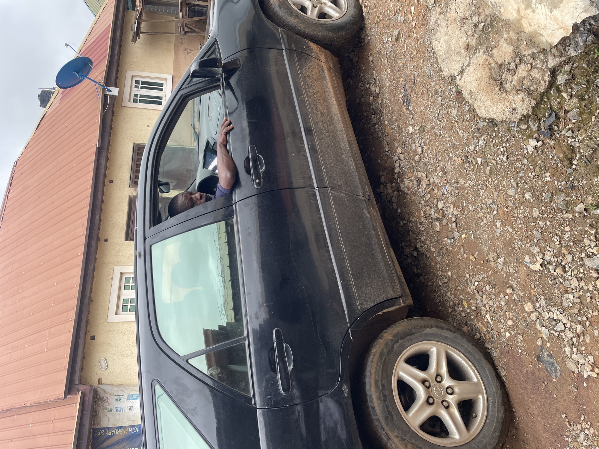 A black SUV is parked on a dirt road in Kurunduma, Nigeria. A Black man is in the driver's seat, with his left arm resting on the open window sill. The car's exterior is dirty, with visible streaks of mud along the lower panels. The surrounding environment is characterized by a light brown, gravelly dirt road, with scattered rocks and debris. In the background, a light yellow building with a reddish-brown corrugated metal roof is visible. A blue satellite dish is mounted on the roof of the building. The sky is overcast.