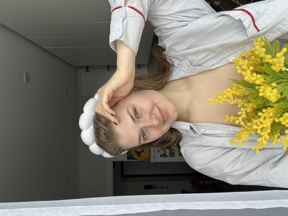 A young adult female is depicted indoors in Nikol'skii, Russia, captured from the chest up. She possesses fair skin, light brown hair, and visible freckles. On her head, she wears a white, segmented headband. Her attire consists of a light-colored, striped collared shirt, unbuttoned, revealing her chest, with red trim visible on the interior of the cuffs and a pocket. Her left hand, with dark nail polish, is raised, fingers resting near her hairline on her forehead. She holds a bouquet of yellow mimosa flowers with green foliage centrally in front of her chest. The subject is looking directly at the camera with a slight smile. The background on the left features a sheer white curtain, an indistinct dark area with a wall-mounted object, a partially visible appliance (possibly a refrigerator) adorned with items, and a yellow circular artwork. The background on the right displays a series of white, flat-panel cabinets or closet doors. The ceiling is white and includes two visible recessed lights.