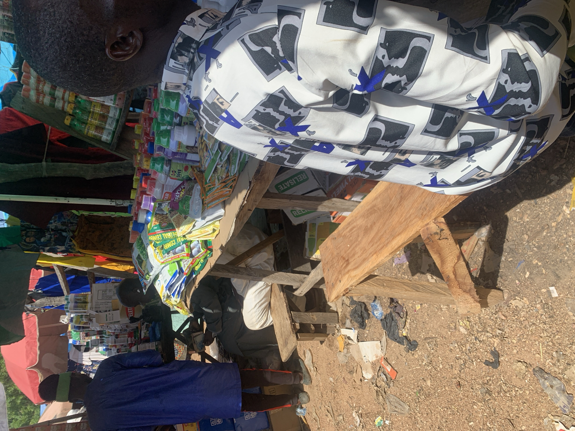 This media file depicts a bustling outdoor market scene in Nigeria. The subjects are primarily two individuals, one wearing a white shirt with a bold black and blue graphic print, and another in a blue robe with a green headband. The man in the white shirt is partially visible, his head and upper body captured as he stands behind a wooden stall laden with various goods. The second individual, wearing the blue robe, is seen from the back, appearing to be a vendor or shopper in the foreground.
The stall itself is filled with an assortment of products, including what appear to be packaged food items, spices, or agricultural supplies, judging by the colorful labels on many of the containers and bags. Some of these items are in small bottles, while others are in larger sacks or boxes. The stall structure is constructed from rough wooden planks. The ground surrounding the stall is dirt and strewn with debris, including plastic bags and discarded materials, suggesting a rustic and perhaps informal market setting.
The overall scene is illuminated by bright sunlight, indicating it is daytime. The shadows cast by the wooden structure and the people suggest the sun is high in the sky. The atmosphere appears busy and transactional, common in a marketplace. There are no strong emotional cues evident in the limited view of the individuals.
Visible text on some of the products includes what appears to be branding and product names. For example, one package prominently displays "GLOBAL COMBI" and another visible text reads "RELISATE". The presence of these labels helps identify some of the goods being sold. The setting cues, such as the merchandise and the rough construction of the stall, strongly point to a market in a developing region, consistent with the provided location context of Nigeria.