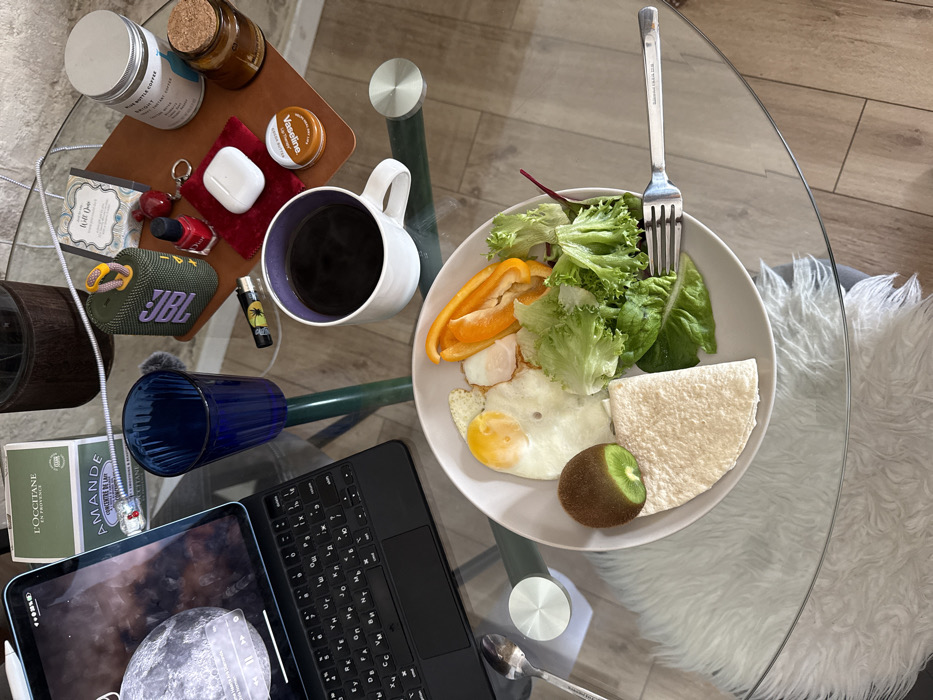 The scene presents a top-down view of a glass table laden with breakfast items and personal belongings, situated in Tel-Aviv, Israel. A white ceramic plate centrally holds two fried eggs, slices of orange bell pepper, a portion of green lettuce, a half-cut kiwi fruit, and a folded light-colored flatbread. To the right of the plate, a white mug contains dark coffee, and an empty blue ribbed drinking glass is positioned above it.
An iPad Air, encased in a dark keyboard folio, rests on the left side of the table, displaying a lunar or planetary image with active media playback controls. Adjacent to the iPad are two L'Occitane en Provence boxes. On the upper right, a dark green JBL portable speaker with an orange carabiner hook is visible. A collection of smaller items includes a small red nail polish bottle, a lighter with a green leaf design, and a white Apple AirPods case, all resting on a red fabric mat. Further right, a silver jar labeled "BLUE BOTTLE COFFEE," a small glass jar with a cork stopper, and a "Vaseline Cocoa Butter" tin are arranged on a brown leather-like mat. A silver spoon and a silver fork (bearing the inscription "BOHMANN") are placed near the plate. The glass table surface reflects a white, fuzzy rug beneath and light-colored flooring in the background. No individuals are present.