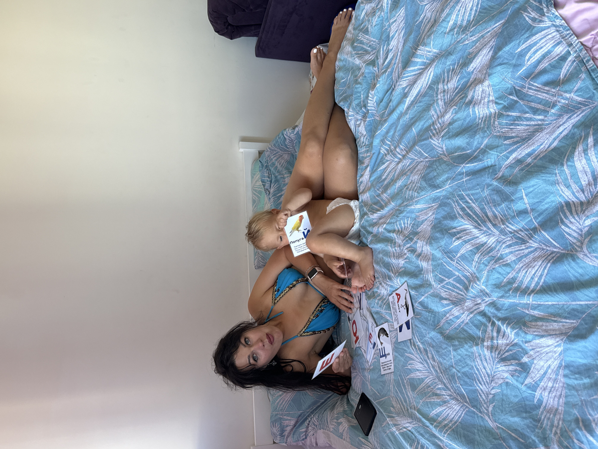 The image shows a woman and a young child lying on a bed with blue and white palm leaf patterned bedding. The woman is wearing a blue bikini with gold embellishments. She has long, dark hair and is looking at the camera with a surprised expression. The child is lying on its side, wearing a diaper, and holding a card with an image of a bird and text in Russian. The woman is also holding cards with letters and images, some of which are visible and contain Russian text. There is a dark object, possibly a phone, on the bed near the woman's feet. The setting appears to be a bedroom, with a plain white wall in the background. A dark purple cushion is visible in the upper left corner, suggesting it is part of a sofa or chair. The lighting suggests it is daytime.