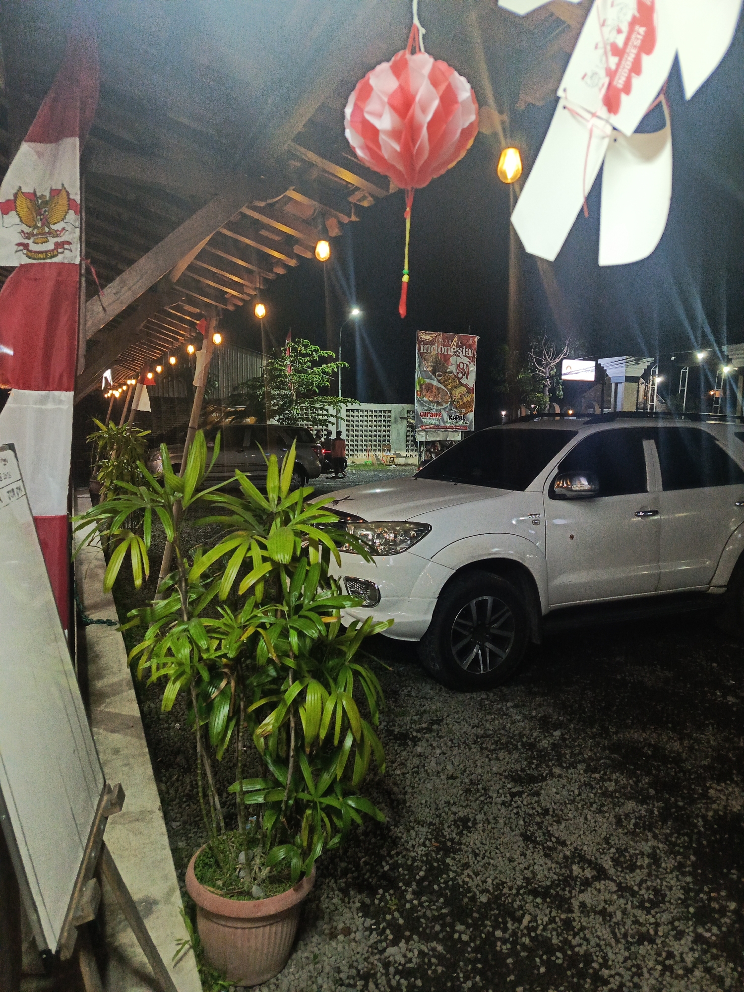An eye-level, medium shot captures a white Toyota Fortuner SUV parked on a gravel surface at night. The vehicle is positioned in front of a building with a wooden roof structure, illuminated by string lights and a red and white paper lantern. A potted plant with lush green foliage sits in the foreground to the left of the vehicle. To the left of the plant, an Indonesian flag with the Garuda Pancasila emblem is displayed. In the background, a banner with Indonesian text and images of food is visible. The overall scene suggests an outdoor dining or event setting in Banguntapan, Indonesia, likely during a celebration given the flag and decorations. No people are clearly visible interacting with the vehicle or surroundings.