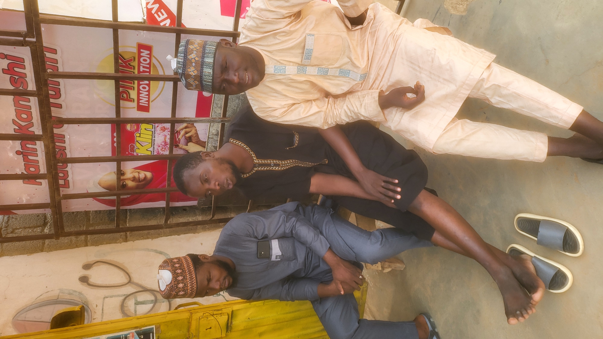 Three men are posed in front of what appears to be a storefront in Jogana, Nigeria. The man at the top is wearing a light orange traditional outfit with patterned embroidery, and a kufi cap. The man in the middle is wearing a black robe with gold-colored embroidery. The man at the bottom is wearing a gray tunic and pants, and a kufi cap. The men are all sitting or reclining. A pair of gray sandals are visible on the ground. Behind them is a barred window or gate, displaying advertisements. On the wall to the left of the men, there are medical symbols painted, including a stethoscope and a doctor's helmet. The ground is light tan.