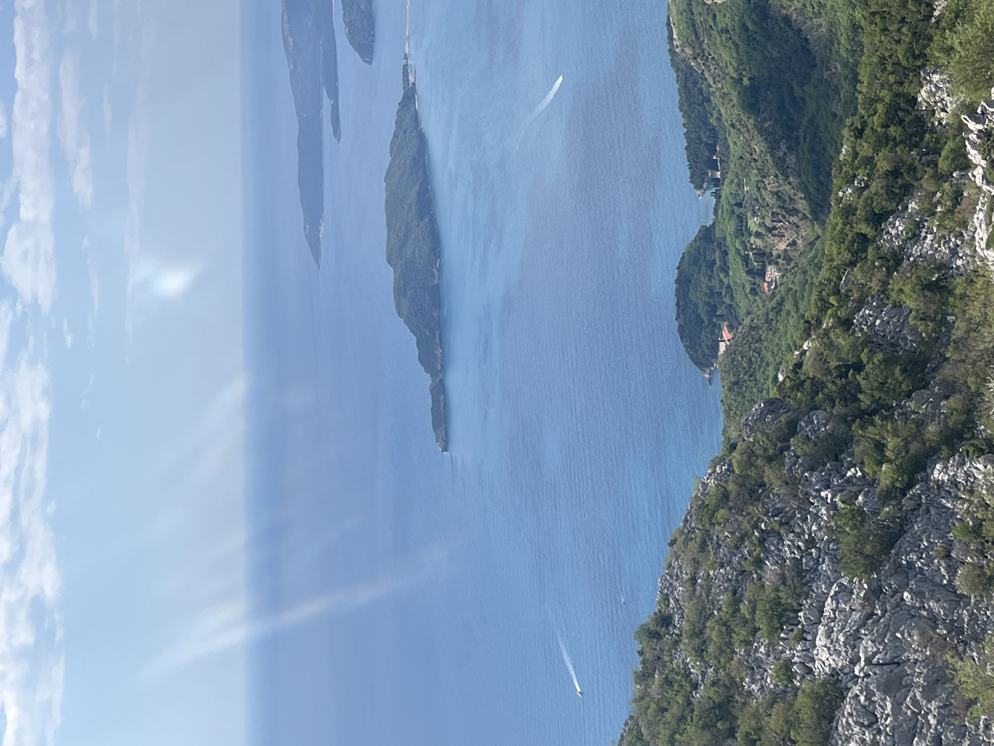The image captures a scenic view from a high vantage point near Boreti, Montenegro. The scene shows several islands stretching into the Adriatic Sea. Turquoise water surrounds the islands. The shoreline includes rugged, rocky terrain covered with dense, green vegetation. Two small boats are visible on the water, leaving white wakes behind them. The sky is partly cloudy.