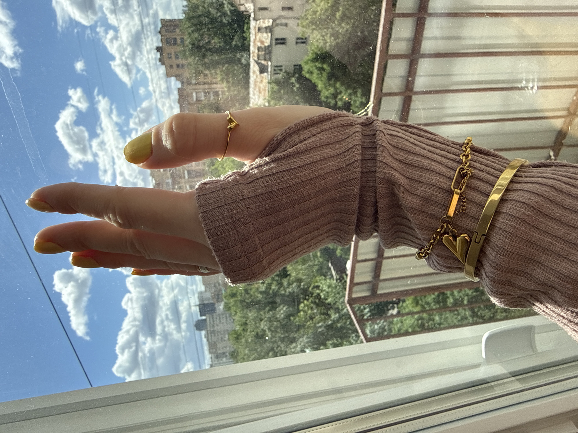 A person's arm, wearing a long-sleeved, ribbed mauve sweater, is extended in front of a window. The person is wearing a gold heart ring on the index finger, and a gold bracelet and bangle on the wrist. The person's nails are painted a pale yellow. Outside the window, the sky is blue with fluffy white clouds. In the distance, there are buildings and trees. The environment appears to be Moscow, Russia, based on the architecture and foliage.