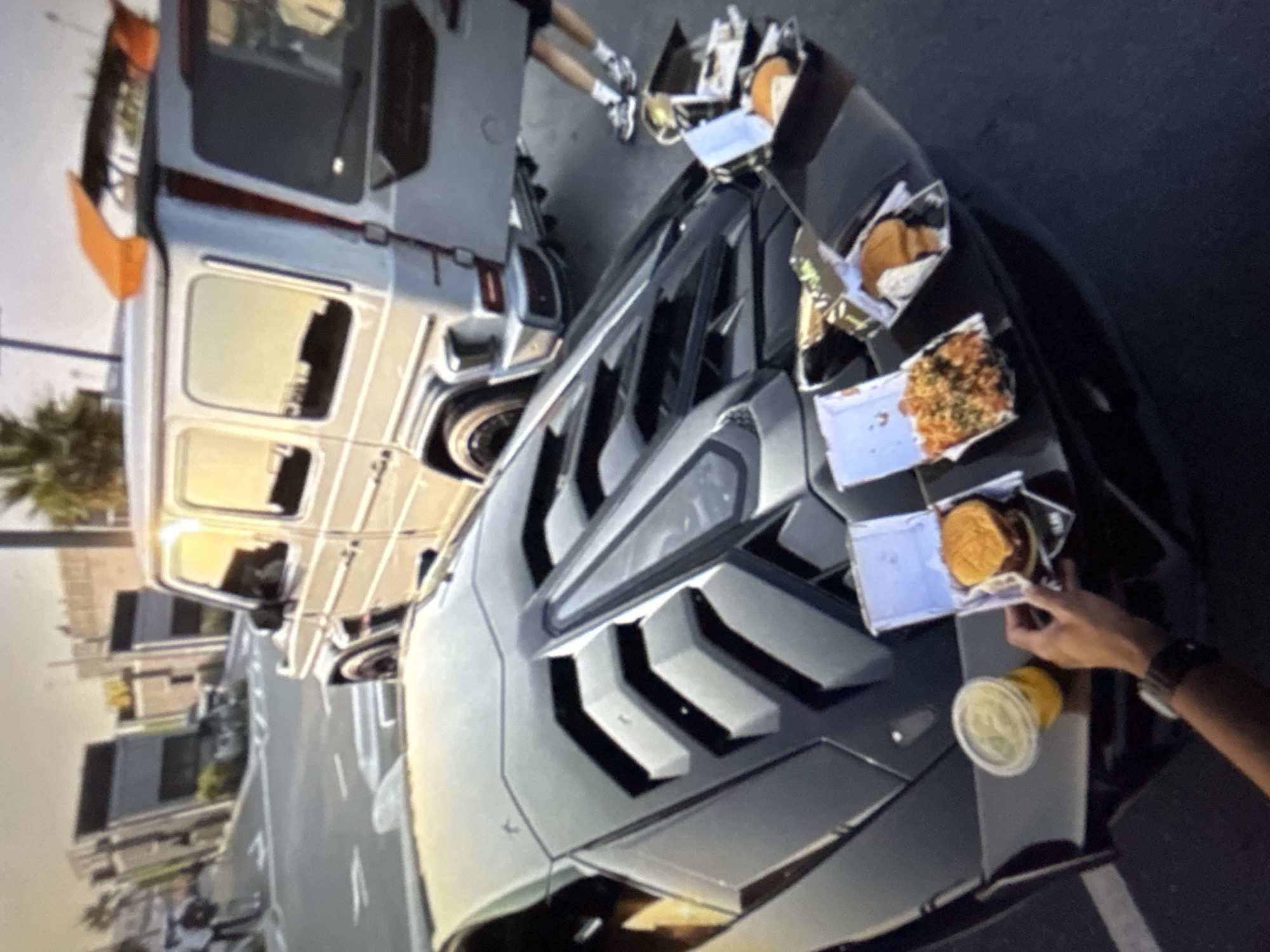 The image depicts an outdoor scene in Burgas, Bulgaria, featuring two luxury vehicles and several takeout food items. In the foreground, a black Lamborghini, identifiable by its distinctive slatted engine cover and aerodynamic design, is parked on an asphalt surface. The vehicle's rear spoiler and engine bay are utilized as a surface for multiple open takeout containers and a drink. One container holds a burger, another contains what appears to be seasoned fries or rice, and at least two other containers hold additional food items, including another burger wrapped in paper. A clear plastic cup with a light-colored beverage is also present. A person's arm and hand are visible in the lower right foreground, holding one of the burger containers; a dark-banded wristwatch is visible on their wrist.
Behind the Lamborghini, a silver Mercedes-Benz G-Wagen (G-Class) is parked, facing away from the viewer. This vehicle features an aftermarket orange rear spoiler. Near the rear of the G-Wagen, the lower legs and feet of a second individual are visible, clad in athletic shoes and shorts. The ground is asphalt with white painted road markings, including arrows. In the background, modern, multi-story buildings with dark facades and prominent window lines are visible. Palm trees are scattered throughout the distant background, and two additional human figures can be seen walking on a sidewalk. The scene is illuminated by natural light, suggesting daytime conditions.