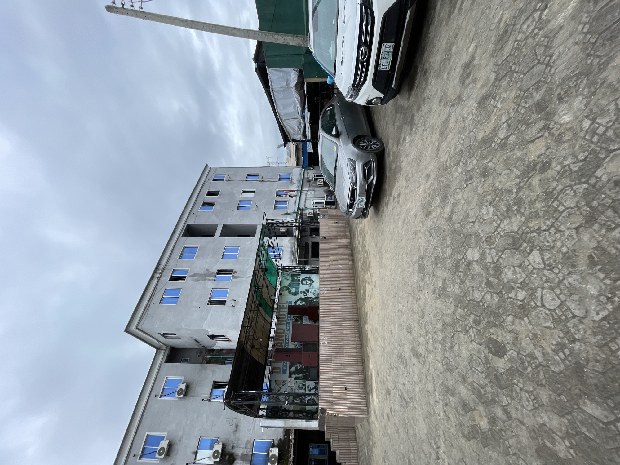 The image shows a multi-story building in Ado, Nigeria. The building is light gray with blue-tinted windows and several air conditioning units. In front of the building is an outdoor stage area with a black canopy and a backdrop featuring portraits. Two cars are parked in the foreground on a paved surface. The sky is overcast.