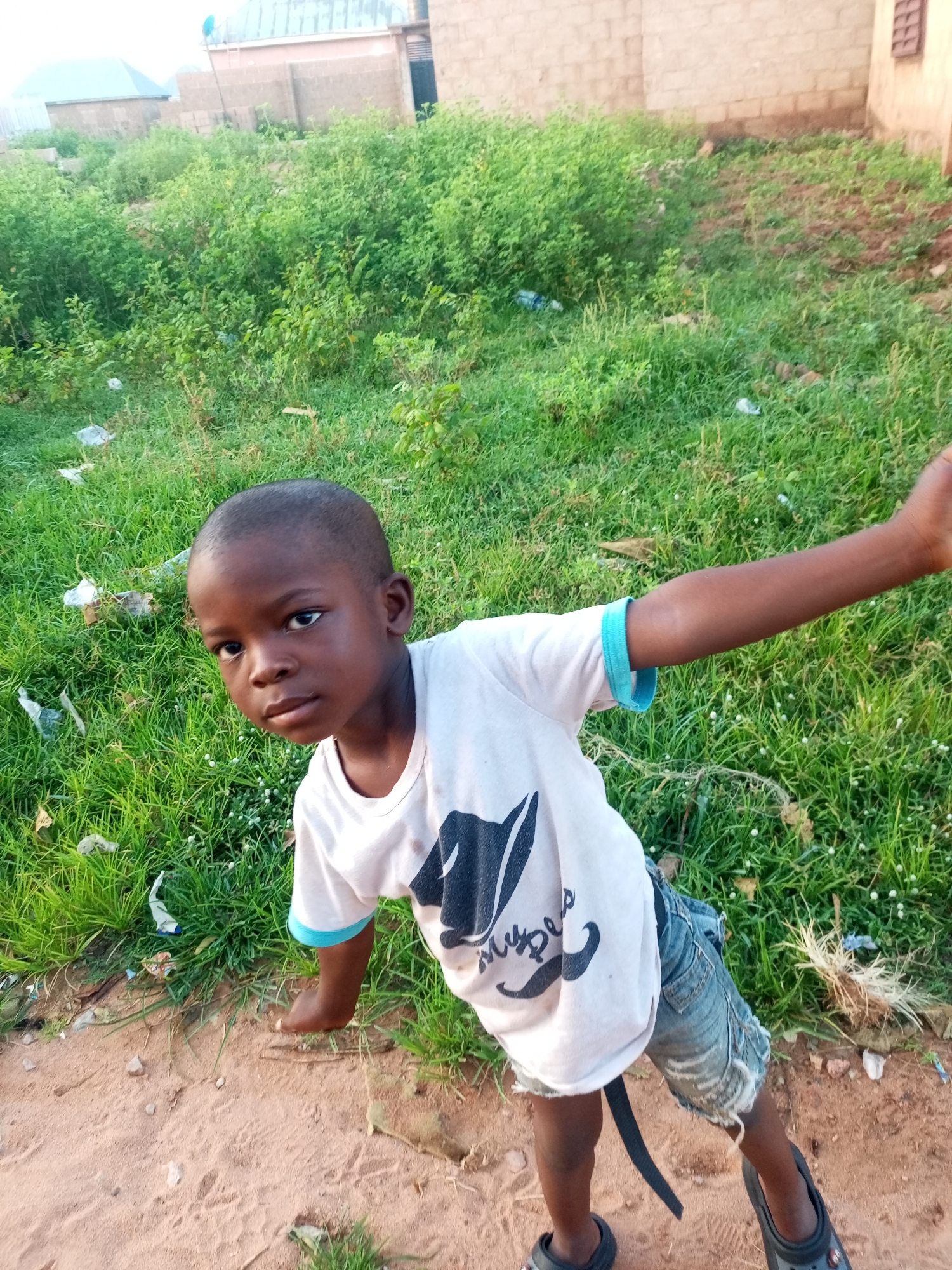 In Tempure, Nigeria, a young boy stands on a dirt path bordering a patch of overgrown green grass. He wears a white t-shirt with a black graphic and blue trim on the sleeves, and ripped denim shorts. His right arm is extended to the right, while his left arm is bent at the elbow, with his hand resting on his leg. The background features a brick wall and some buildings. The scene appears to be outdoors, with natural light illuminating the boy and his surroundings.