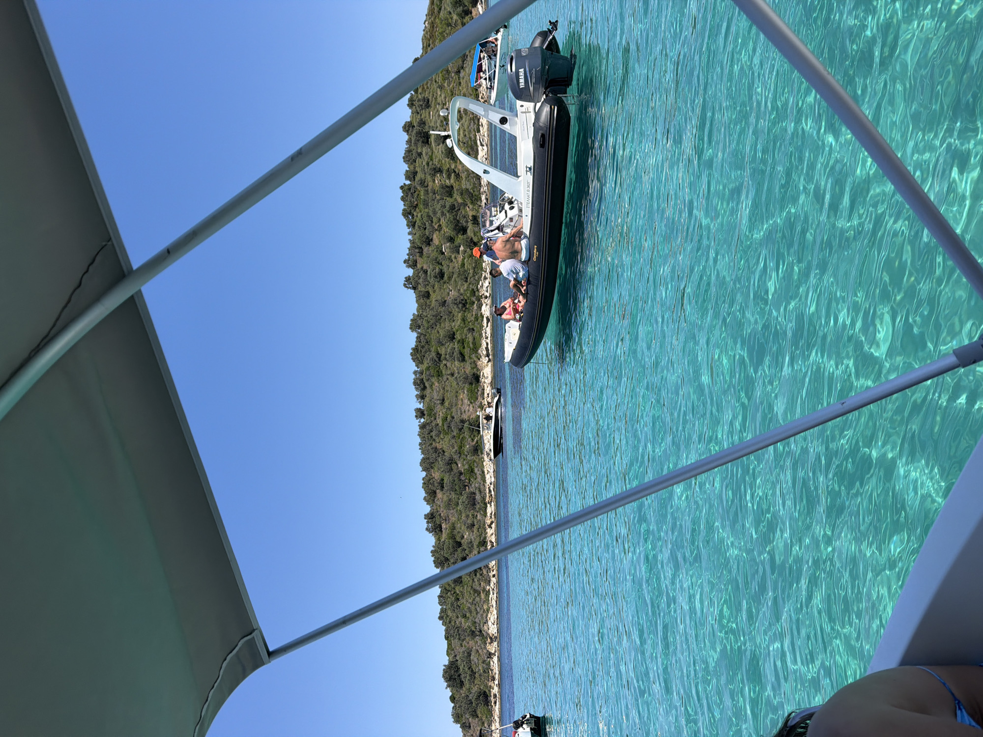 The image shows a bright, sunny day with clear turquoise water. Several boats are present, including a rigid inflatable boat with a Yamaha engine carrying four people. The boat is dark with a white interior. In the background, there is a rocky, tree-covered island. The foreground shows part of another boat with a canopy, suggesting the photo was taken from another vessel. The scene depicts a leisurely outing on the water.