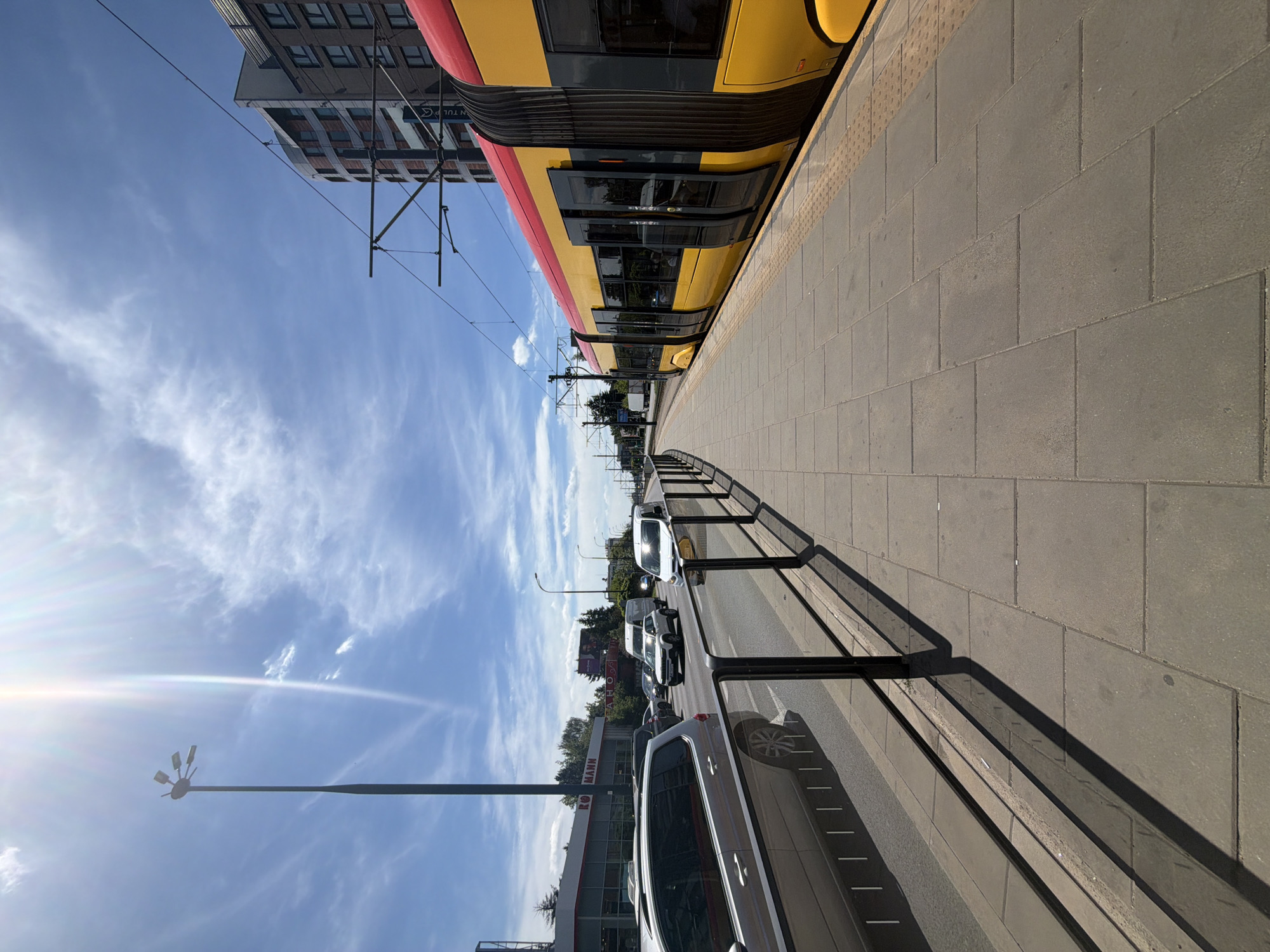 An outdoor, eye-level, full shot captures a sunny day in a city with a bright blue sky and some white clouds. In the foreground, a wide sidewalk paved with square gray tiles runs alongside a tram platform. A yellow and red tram is stopped at the platform, its doors open. A black metal fence separates the sidewalk from a street with multiple lanes of traffic. Several cars, including a silver minivan and a white van, are driving on the street. To the left of the street, a building with a red and white sign that reads "ROMANN" is visible, along with trees and streetlights. Further in the background, a tall, modern building stands. The sun is shining brightly, casting long shadows on the sidewalk and street.
