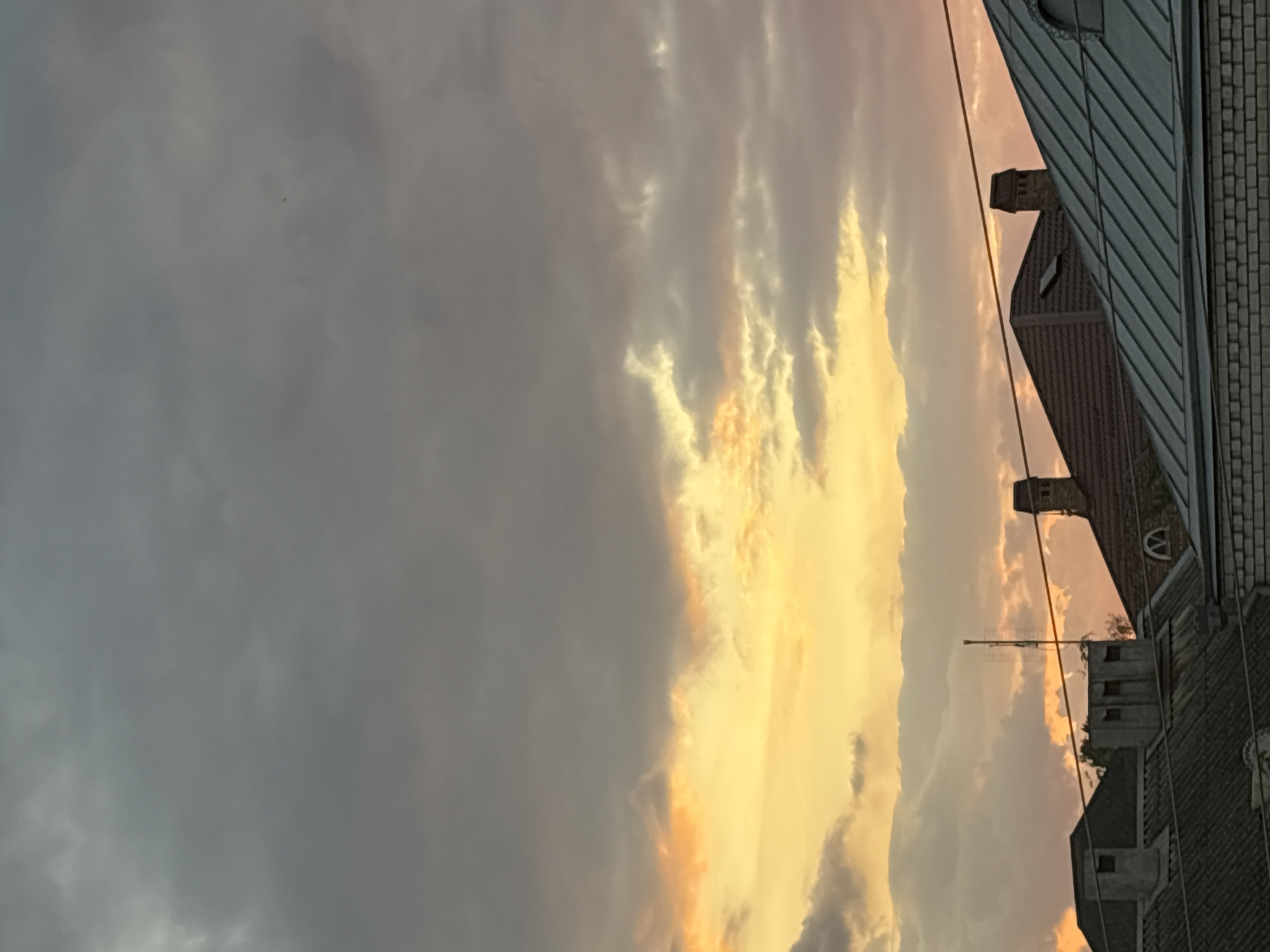 The image captures a dusky sky with scattered clouds illuminated by the setting sun, casting a warm orange and yellow glow. The foreground features the rooftops of several houses with chimneys and antennas. The architectural style suggests an urban or suburban setting. The specific location in Georgiyevsk, Russia, is not visually evident from the image itself. There are no people or discernible actions taking place. The scene is static and primarily focuses on the interplay of light and shadow on the buildings and clouds.