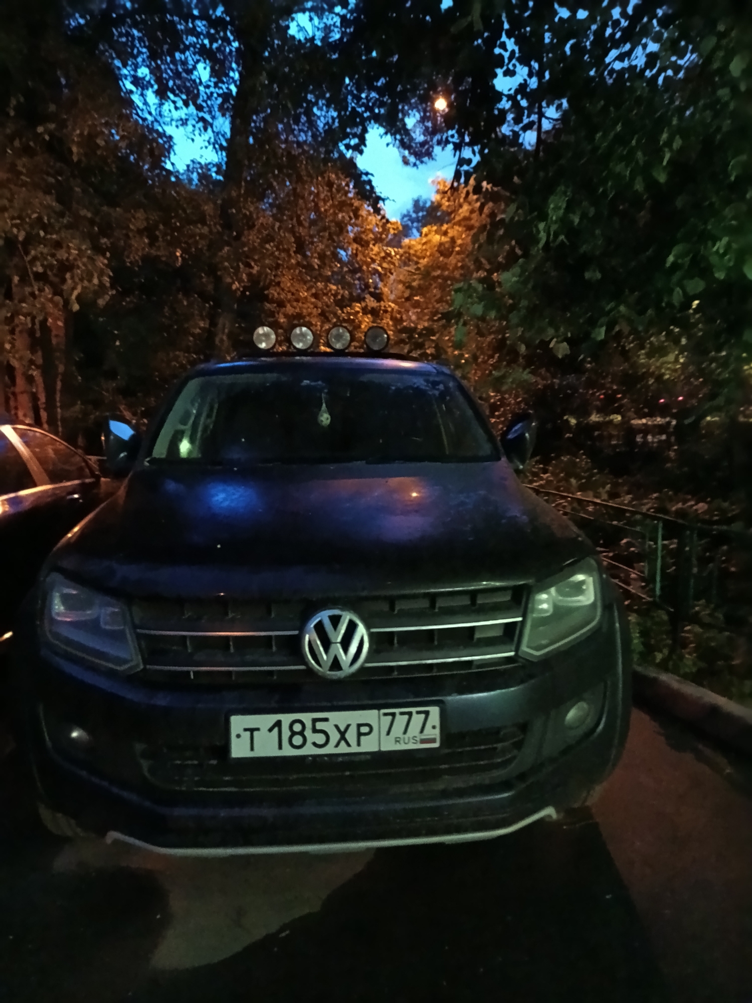The image shows the front of a dark-colored Volkswagen Amarok parked on a street. The car has additional lights mounted on its roof. The license plate reads "T 185 XP 777 RUS". The surrounding environment appears to be a street in Moscow at dusk or night, indicated by the lighting, trees, and vegetation.