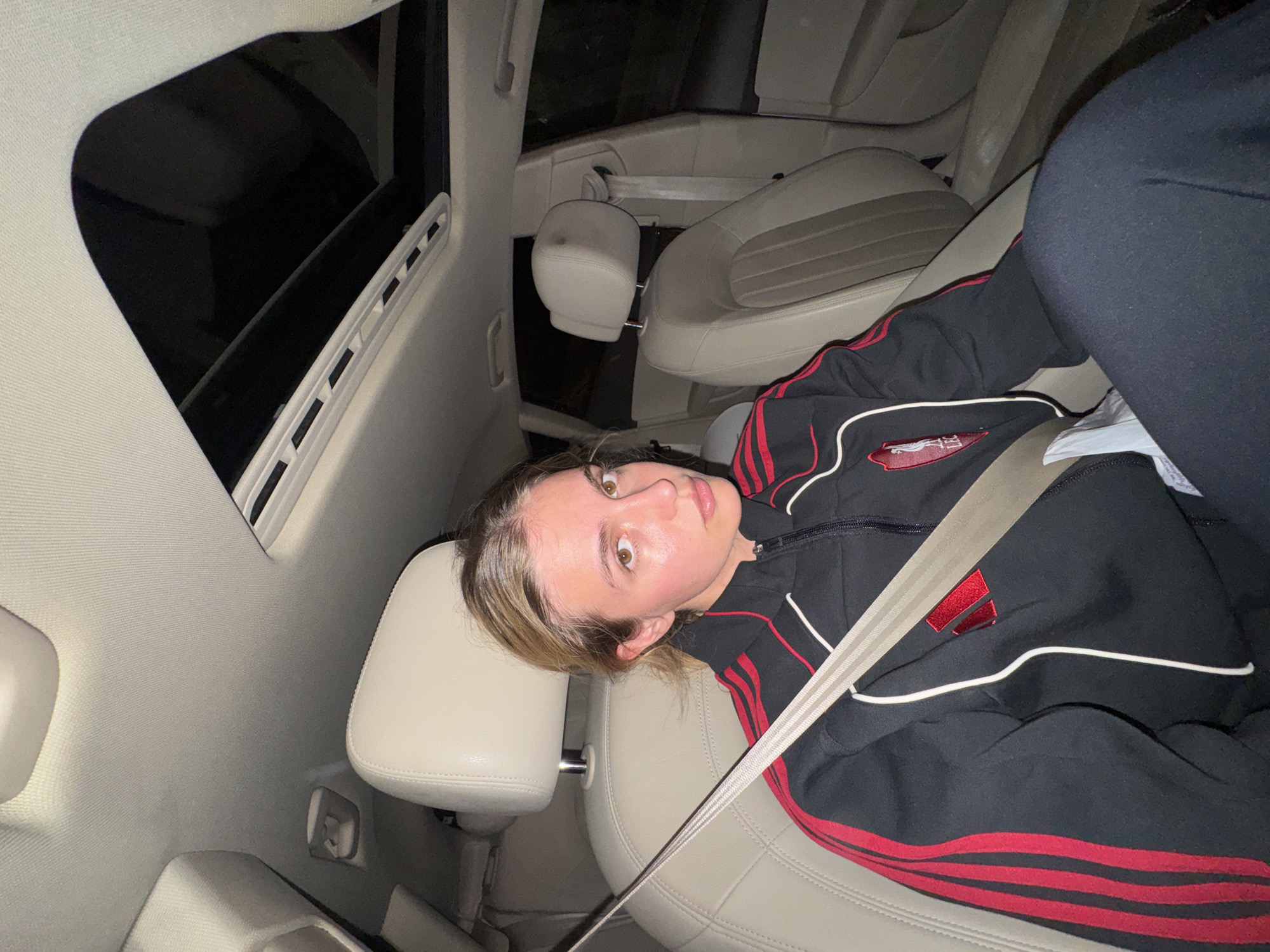 The image captures a young person inside a car, looking directly at the camera. The subject has light brown hair and brown eyes, and is wearing a black tracksuit jacket adorned with red stripes on the sleeves and white piping. A patch on the left side of the jacket's chest features a crest that resembles the Liverpool FC "Liver Bird" symbol. The person also appears to be wearing black trousers or leggings.
They are seated in what looks like the front passenger seat, secured by a light-colored seatbelt. The car's interior is characterized by light-colored (possibly beige or light grey) leather seats and headrests. A sunroof is visible in the top left, revealing dark conditions outside, suggesting it is nighttime. Other parts of the car's headliner, a grab handle, and a glimpse of the rear seats are also discernible.
The photograph is taken from a perspective that makes the person appear to be lying on their side due to a 90-degree clockwise rotation. The person maintains a neutral and direct gaze towards the camera. The overall setting is the interior of a vehicle.