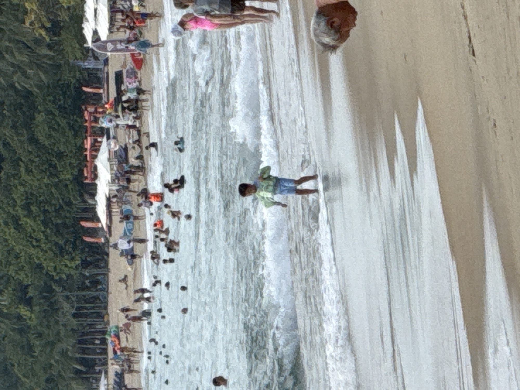 The image depicts a crowded beach scene in Nha Trang, Vietnam, with numerous people enjoying the water and the sand. In the foreground, waves are breaking on the shore, and a young child wearing a life vest and blue shorts stands near the water's edge. Further back, more people are visible in the ocean, some with what appear to be flotation devices. The background reveals a busy shoreline with many beach umbrellas, lounge chairs, and palm trees lining the beach. Several individuals are walking along the sand, and some are seated under umbrellas. The overall atmosphere is one of a lively and popular tourist destination.