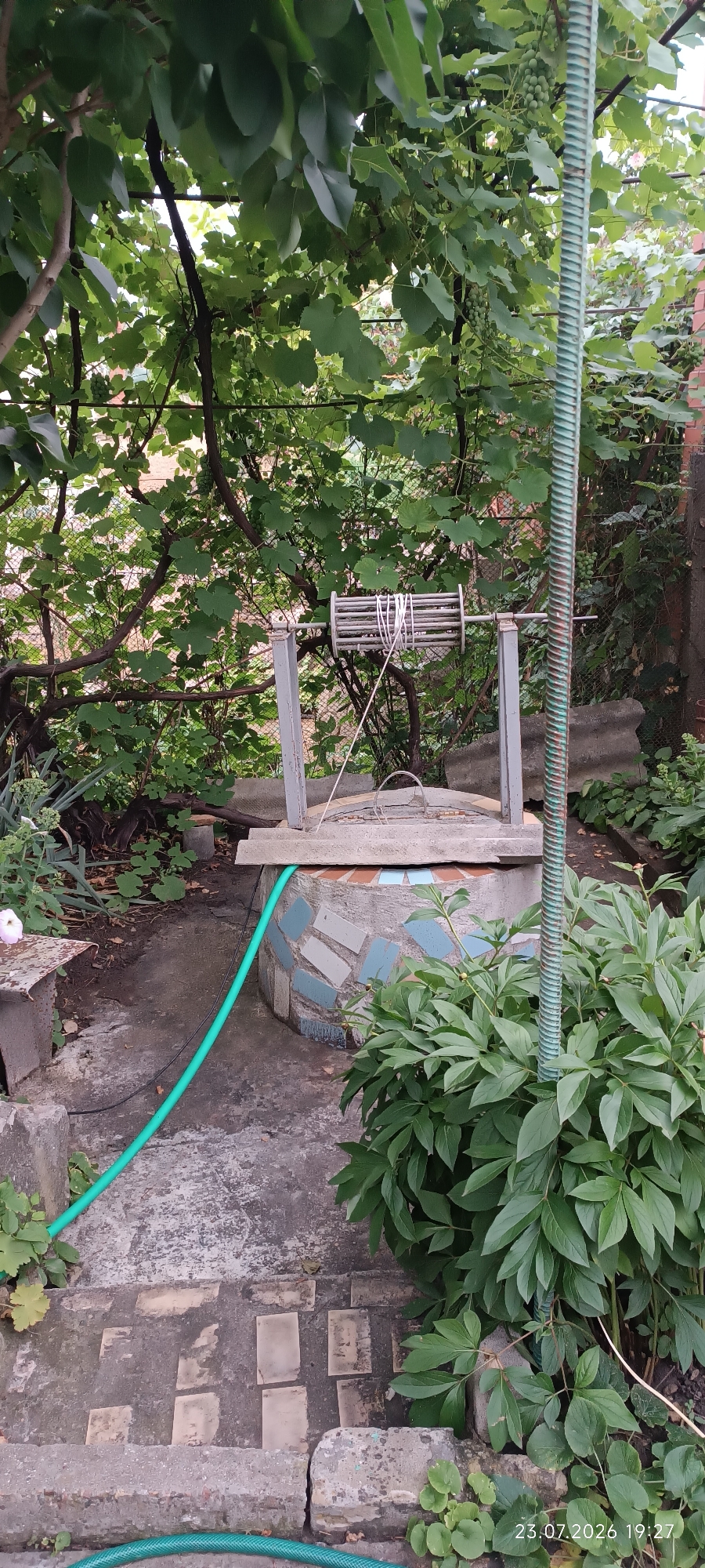 In Bohnatove, Ukraine, a well is pictured in an outdoor setting. The well's base is circular and decorated with mosaic tiles in blue and light gray. A wooden plank sits on top of the well, with a metal mechanism for lowering a bucket. A green hose snakes from the well towards the foreground. The well is surrounded by concrete stairs and paving stones. Lush green foliage, including leaves from trees and bushes, frames the scene. A tall, ribbed green pole stands to the right of the well. The overall environment suggests a garden or yard.