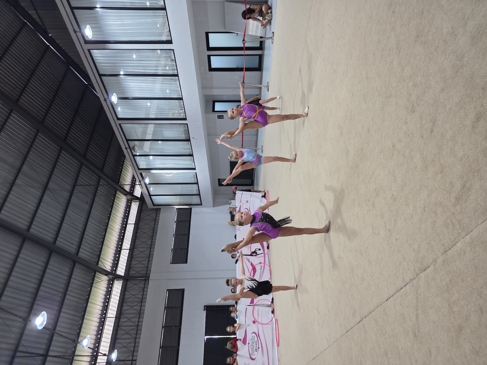 The image shows a group of young female gymnasts performing synchronized splits on a beige floor, likely during a competition or practice session, with a backdrop of promotional banners and an audience. The setting appears to be an indoor sports hall or gymnasium with a high ceiling. North Kuta, Indonesia, is not evident from the image.