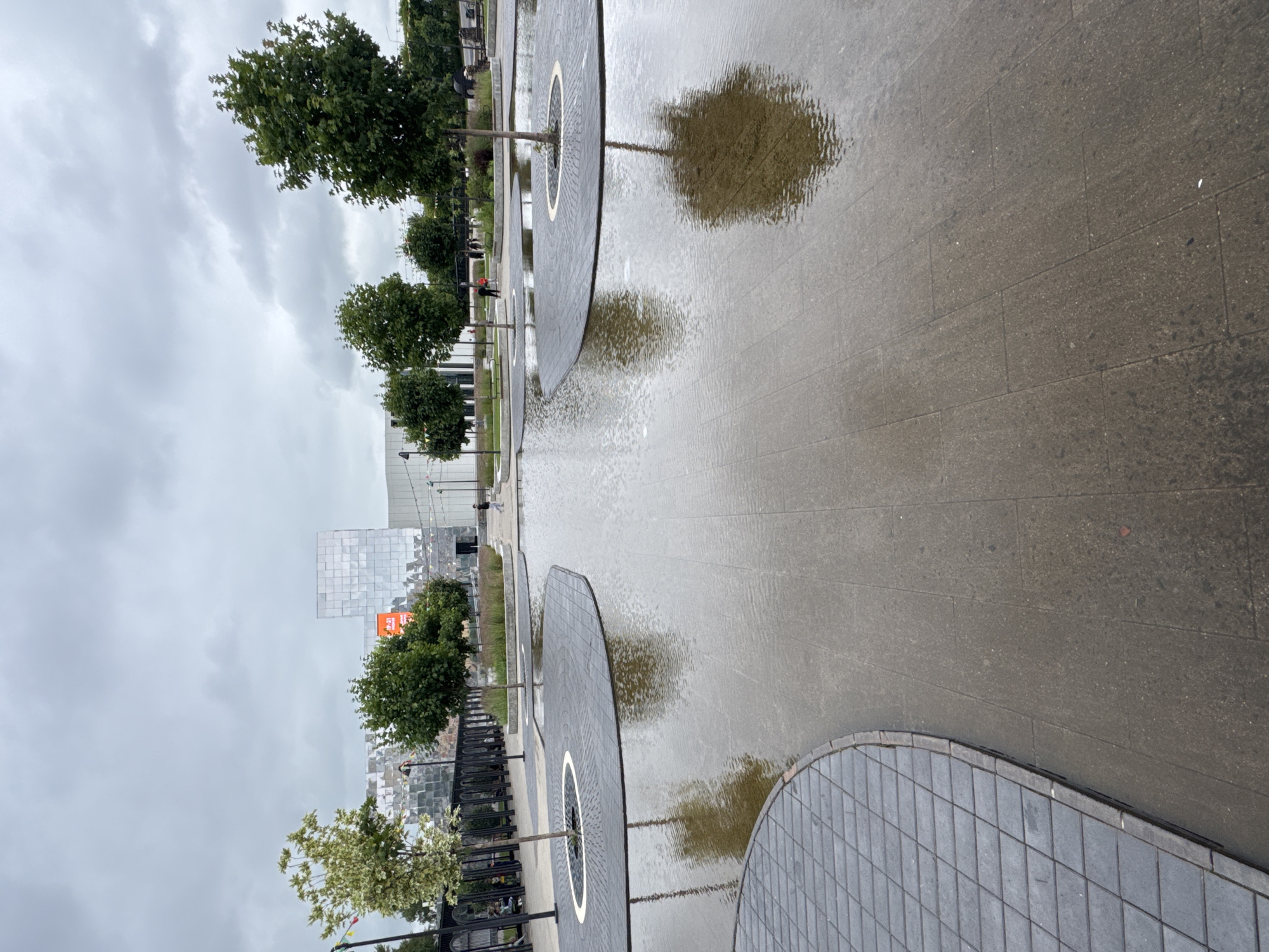 The image depicts an outdoor public space with a shallow water feature. Paved areas with large, grey rectangular tiles are visible, as well as curved sections of smaller, dark grey diamond-shaped tiles. Several young trees are planted on raised circular islands made of dark grey tiles with a white ring around the base of each tree.
In the background, a modern building with a reflective, faceted facade stands. A large orange graphic or advertisement is visible on this building. The sky is overcast with heavy grey clouds. A few people are walking in the distance, and one person is visible in the foreground on the right. The overall impression is of a contemporary urban plaza, possibly a public park or a pedestrian area adjacent to a building. The scene is consistent with the design and aesthetic of the Nizhny Novgorod Stadium area, a notable landmark in the city.