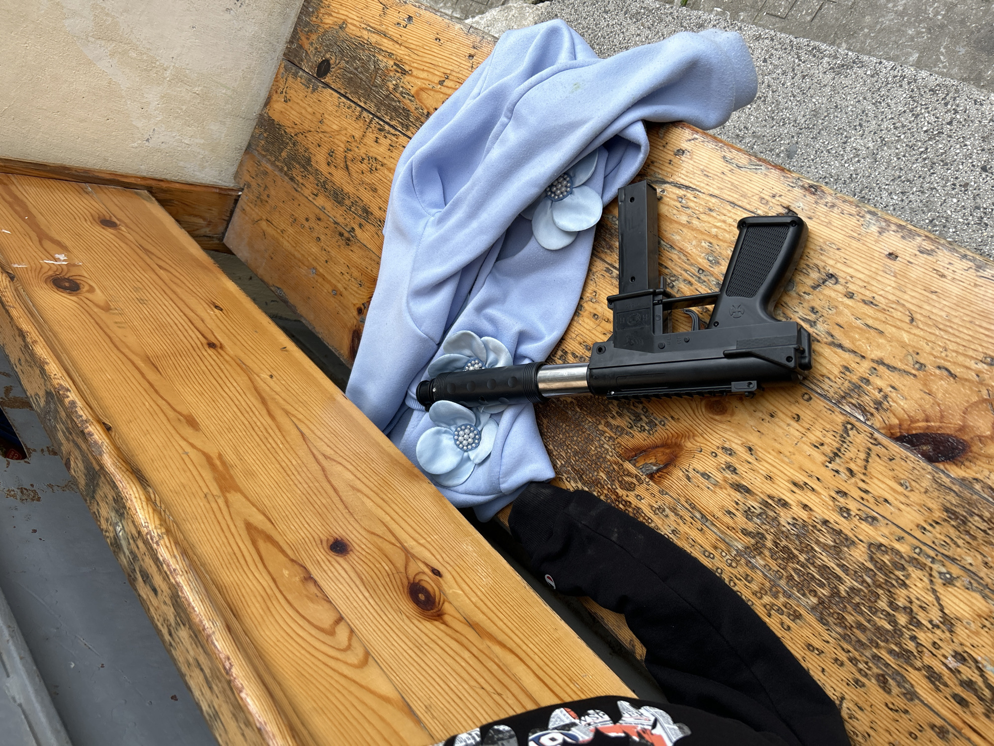 The image shows a weathered wooden bench situated near a concrete sidewalk. Scattered on the bench are clothing items: a light blue sweater adorned with flower embellishments, a black hoodie, and a black firearm with an attached magazine. The bench appears to be positioned outdoors, and there are no visible people or indications pointing to a specific location in Plovdiv, Bulgaria. The scene presents a collection of unrelated objects resting on a park bench.