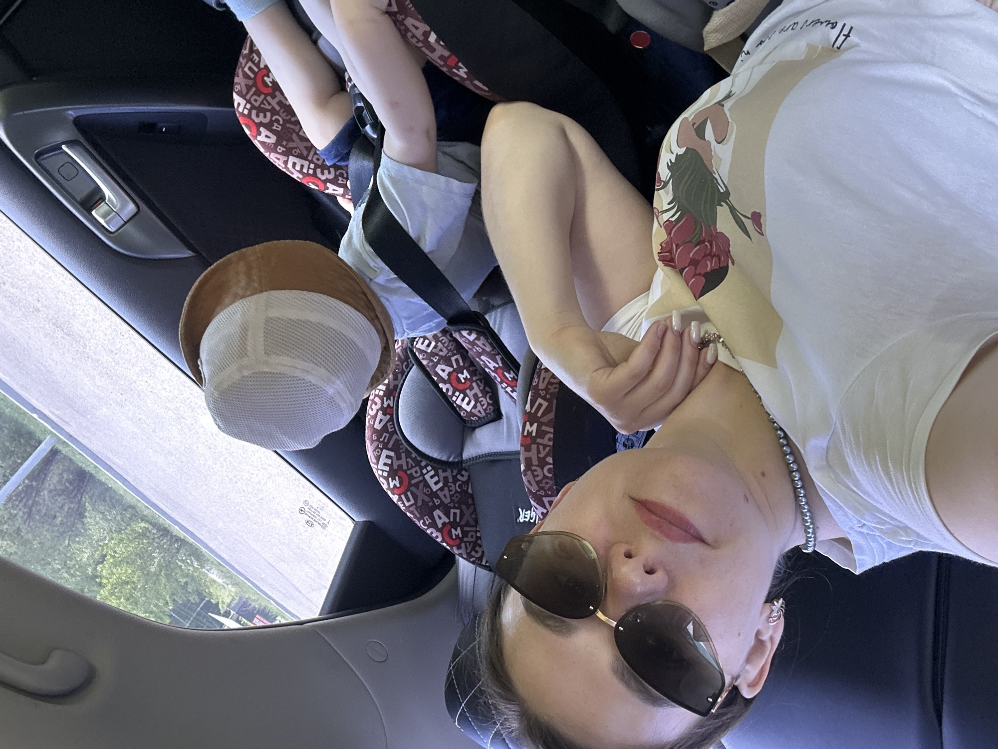The image is an interior shot of a car. A woman with sunglasses is visible in the foreground, partially obscuring the view. She is wearing a white t-shirt with a floral design on the shoulder and a pearl necklace. A child is strapped into a car seat behind her. A tan and white hat sits atop the car seat. Outside the car window, trees and a building are visible. The presence of a KIA logo on the windshield and the car's interior design indicate the make of the car. OCR results include 'EUX', 'M', 'Σ', 'la', '3-', 'AM', 'ΑΠ', 'EH', 'PM', '6', 'PEA', 'GER', and 'flowers are n'. It is not possible to confirm that this photo was taken in the city of Chaikovsky, Russia.