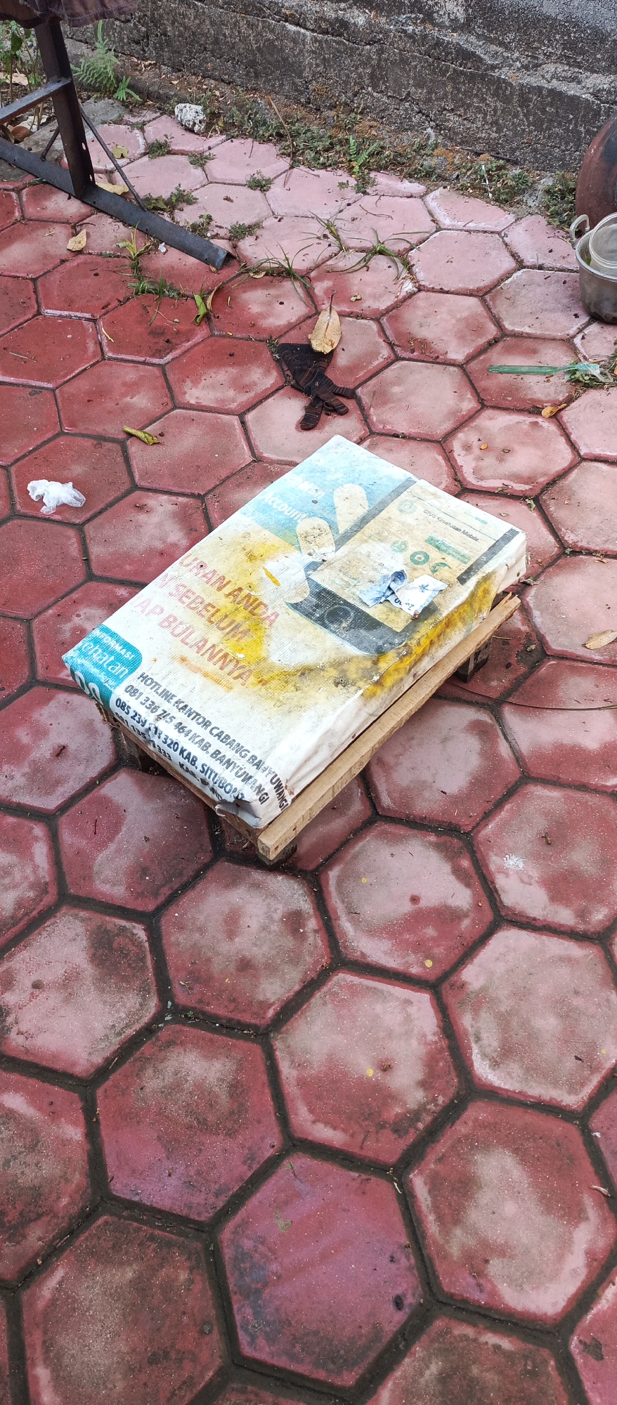 The image depicts a flat, rectangular object, possibly a mattress or a wrapped package, resting on a red, hexagonal-tiled surface. The object is covered in a white material with blue and yellow text and graphics. The text appears to be in Indonesian, mentioning "HOTLINE KANTOR CABANG" and phone numbers. The object is supported by two wooden planks. The tiled area shows signs of wear, with dirt and fallen leaves scattered around. In the background, a concrete wall and some vegetation are visible, along with a metal table leg. A crumpled white piece of paper and a dark, glove-like object lie on the tiles near the object. The scene appears to be outdoors, possibly in a yard or a public space in Arjasa, Indonesia, given the Indonesian text. The lighting suggests it is daytime.