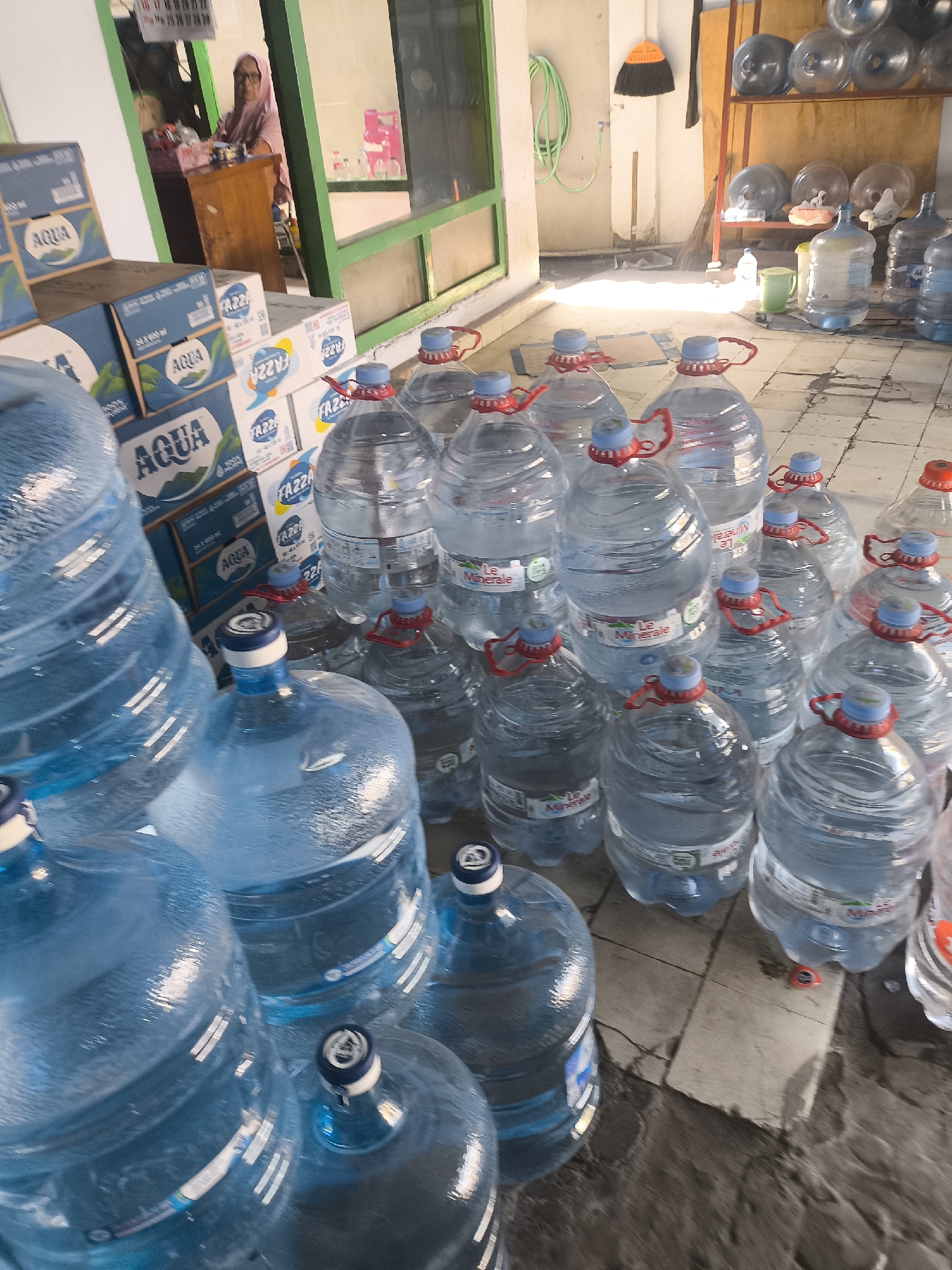 The image depicts a storage area, possibly a shop or warehouse, filled with large plastic bottles of drinking water. The bottles are stacked and arranged in rows on a tiled floor. Several cardboard boxes of bottled water, labeled "AQUA" and "FAZZA," are visible in the background. In the far background, a woman wearing a pink headscarf is seated behind a wooden counter, suggesting a retail or service environment. The lighting suggests it is daytime, with sunlight streaming in from the right. The overall scene is one of bulk water storage and distribution.