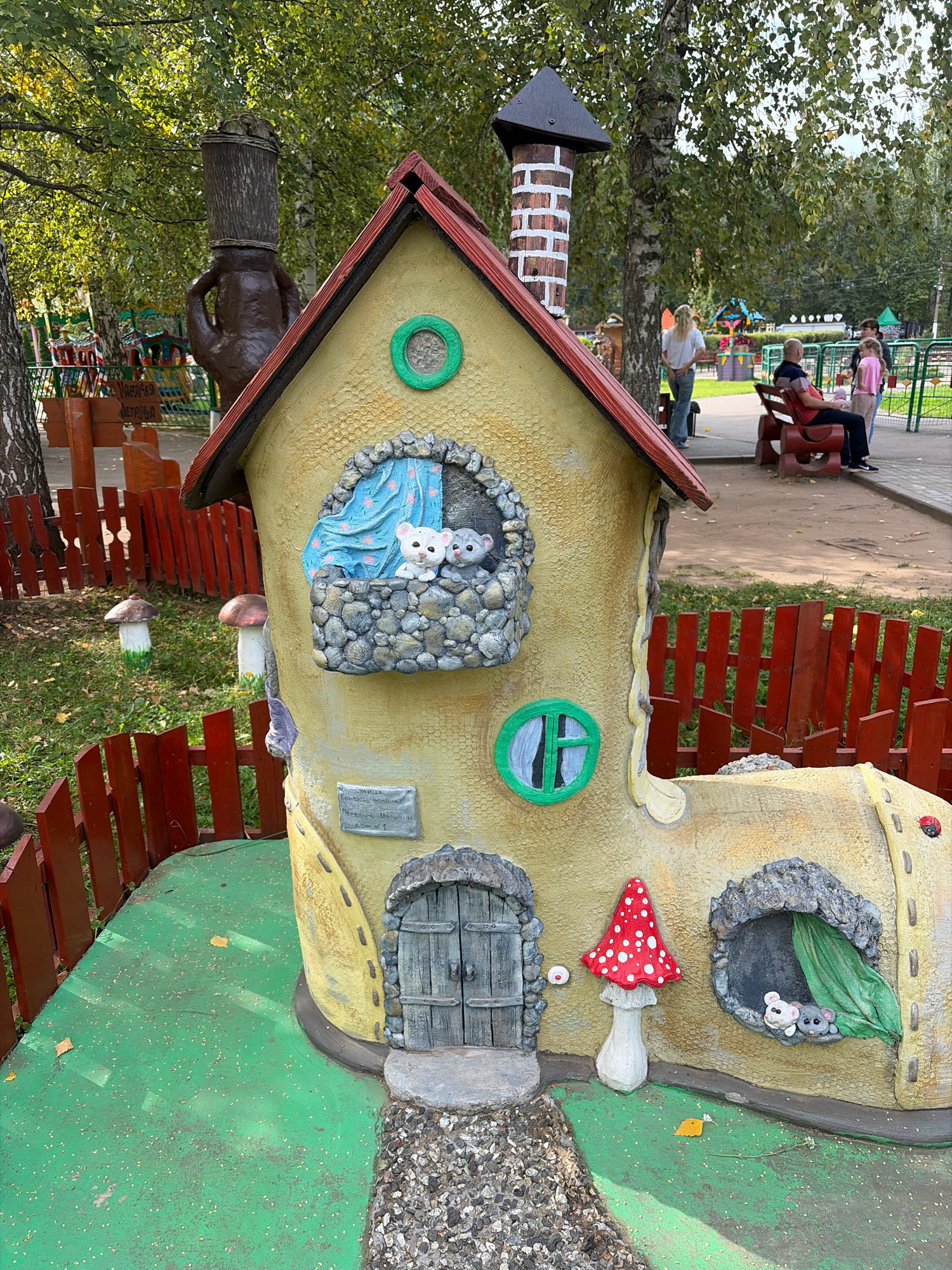 The image shows a whimsical, boot-shaped house sculpture located in Chaikovsky, Russia. The sculpture is painted yellow with a red roof and features a brick chimney. Two small white mice figures are visible in an upstairs window, and two grey mice figures are in a lower window. A red and white spotted mushroom sculpture stands beside the house. A red wooden fence encloses the immediate area around the sculpture. In the background, several people are seen near benches and playground equipment, suggesting this is a public park or playground. Trees with green foliage are present, and the ground is a combination of green artificial turf and dirt pathways. A large, dark brown sculpture of a figure with a wooden bucket for a head is partially visible on the left side of the image.