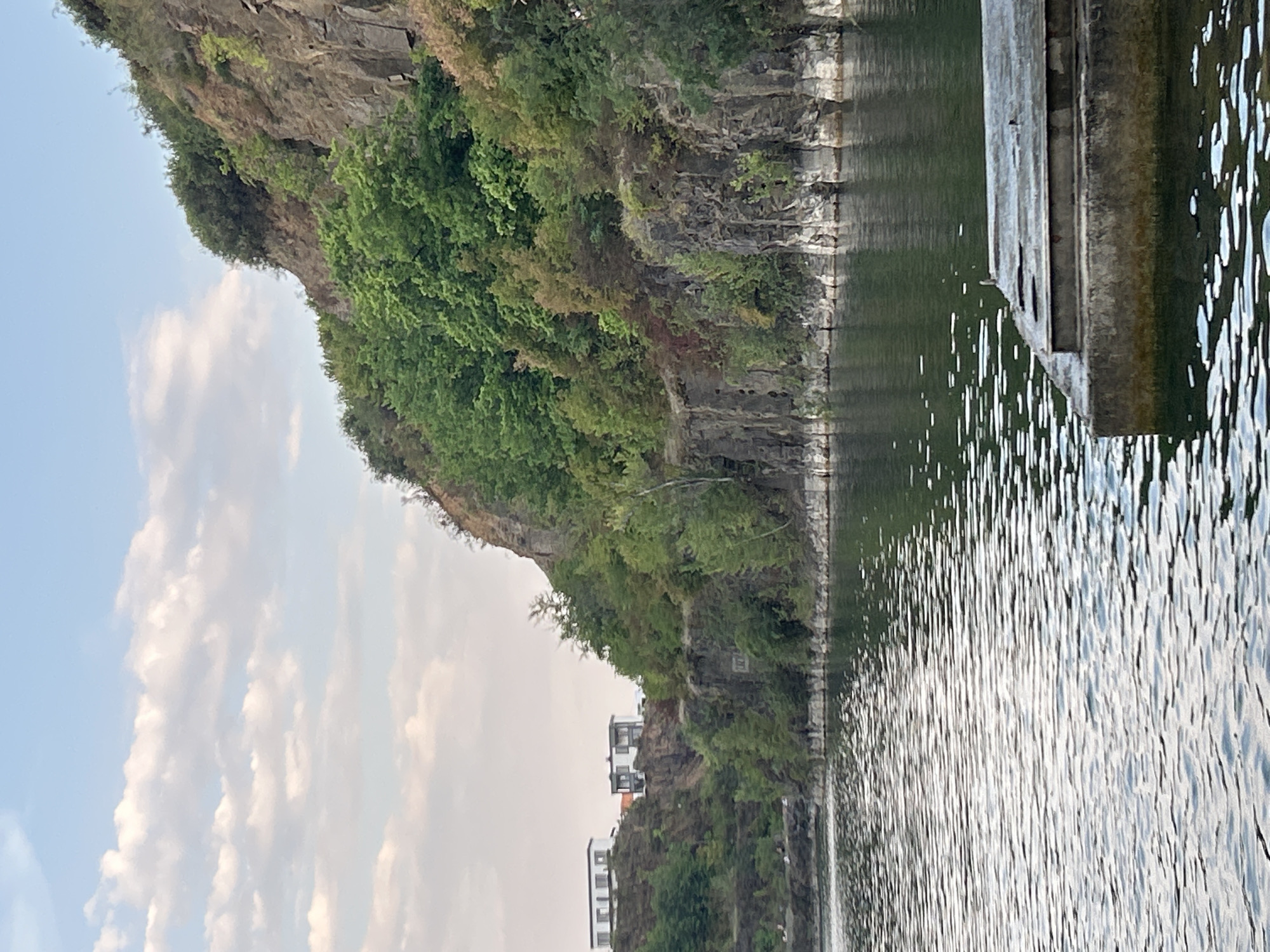 The image depicts a waterfront scene with a rocky, tree-covered cliff face rising from the water's edge. In the distance, on the higher ground of the cliff, are two modern white houses. The water in the foreground is rippled, and a concrete structure, possibly a pier or dock, juts into the frame from the lower right. The sky above is a pale blue with scattered white clouds. The scene is likely a recreational area along a body of water, possibly a quarry that has been transformed into a lake, a common feature in Uzhhorod, Ukraine, where such geological formations and residential developments near water are present.