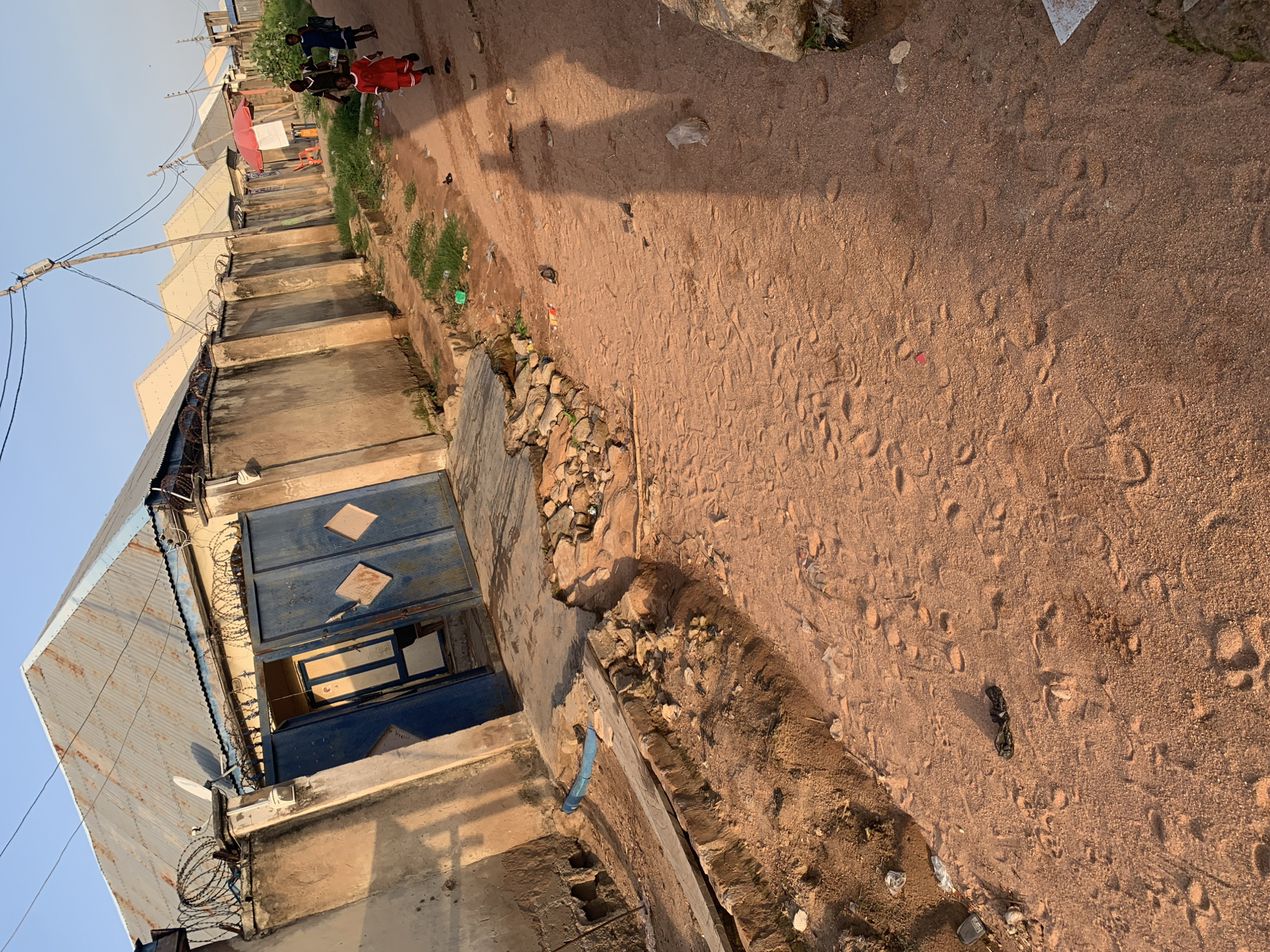 This image depicts an outdoor scene in Jalingo, Nigeria, featuring a dirt path in front of a row of buildings. The buildings have corrugated metal roofs and concrete walls, some topped with barbed wire. A large blue gate, partially open, is visible on the left. Several individuals, including children, are walking or running on the path. The ground is covered in sand and footprints, with scattered debris such as plastic bags and small stones. The sun is casting long shadows, indicating it is likely late afternoon or early morning. Electrical poles and wires are present in the background.