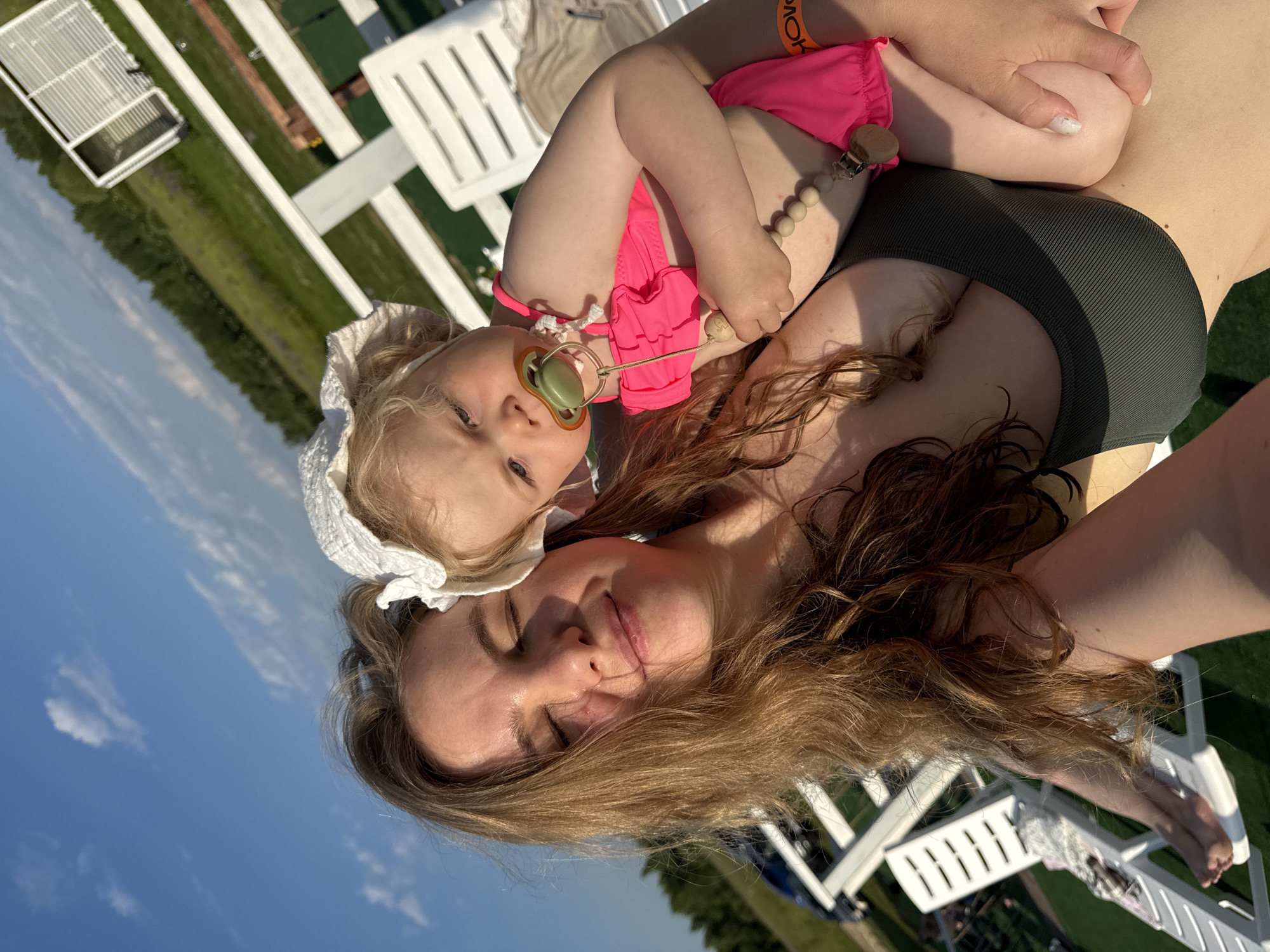 A woman with long, wavy brown hair and closed eyes smiles as she holds a baby. The baby, wearing a white bonnet and a bright pink swimsuit, has a pacifier in its mouth. They are outdoors, with a bright blue sky and some clouds visible. In the background, there are white lounge chairs, a white railing, and trees. The location is identified as Угор, Russia.