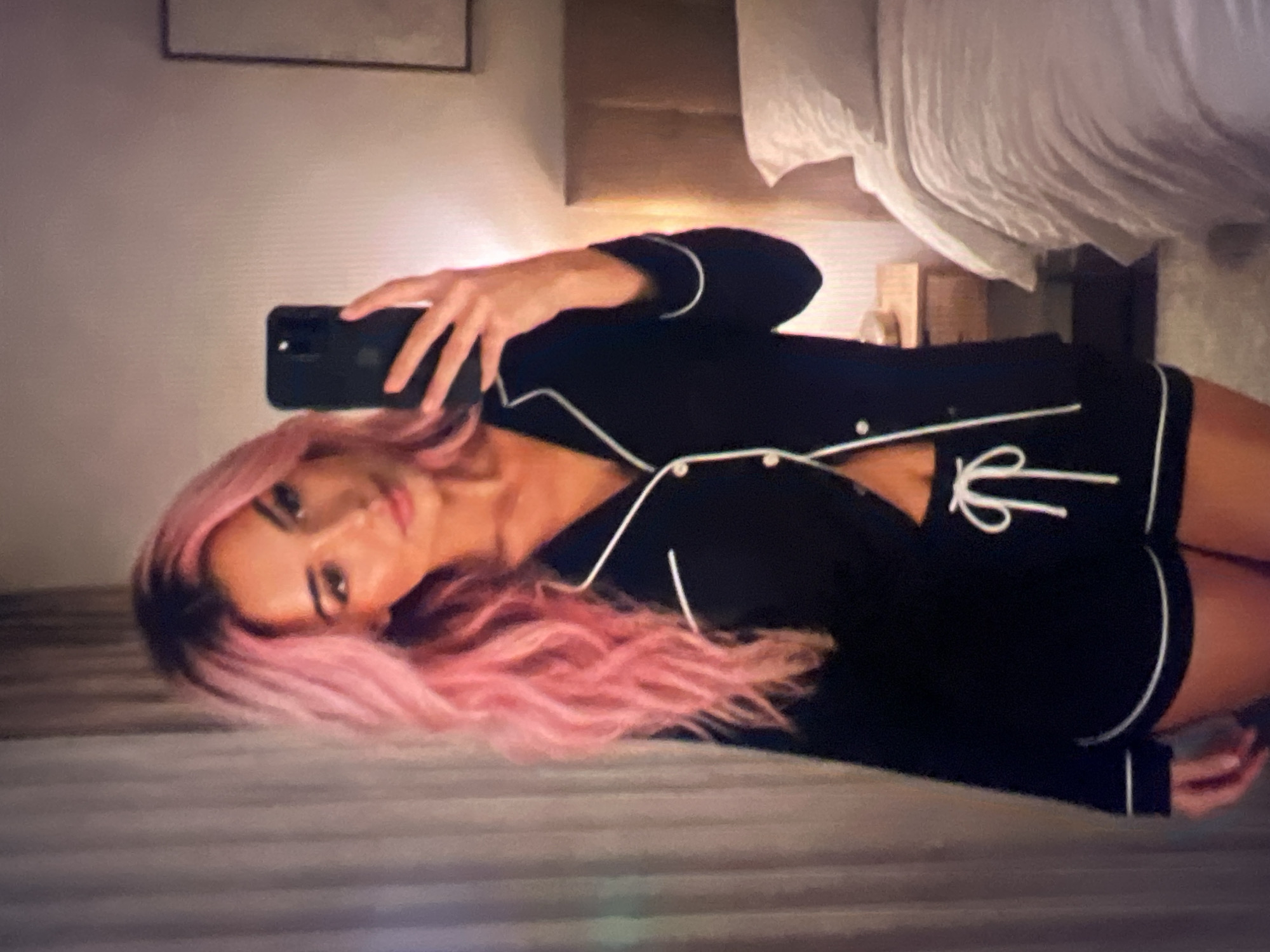 A woman with long, wavy pink hair is taking a mirror selfie in what appears to be a bedroom. She is wearing black pajamas with white piping, consisting of a button-up top and shorts with a drawstring. She is holding a black smartphone in her right hand, angled to capture her reflection. In the background, a bed with white bedding is visible, along with a nightstand. Curtains hang on the left side of the frame.