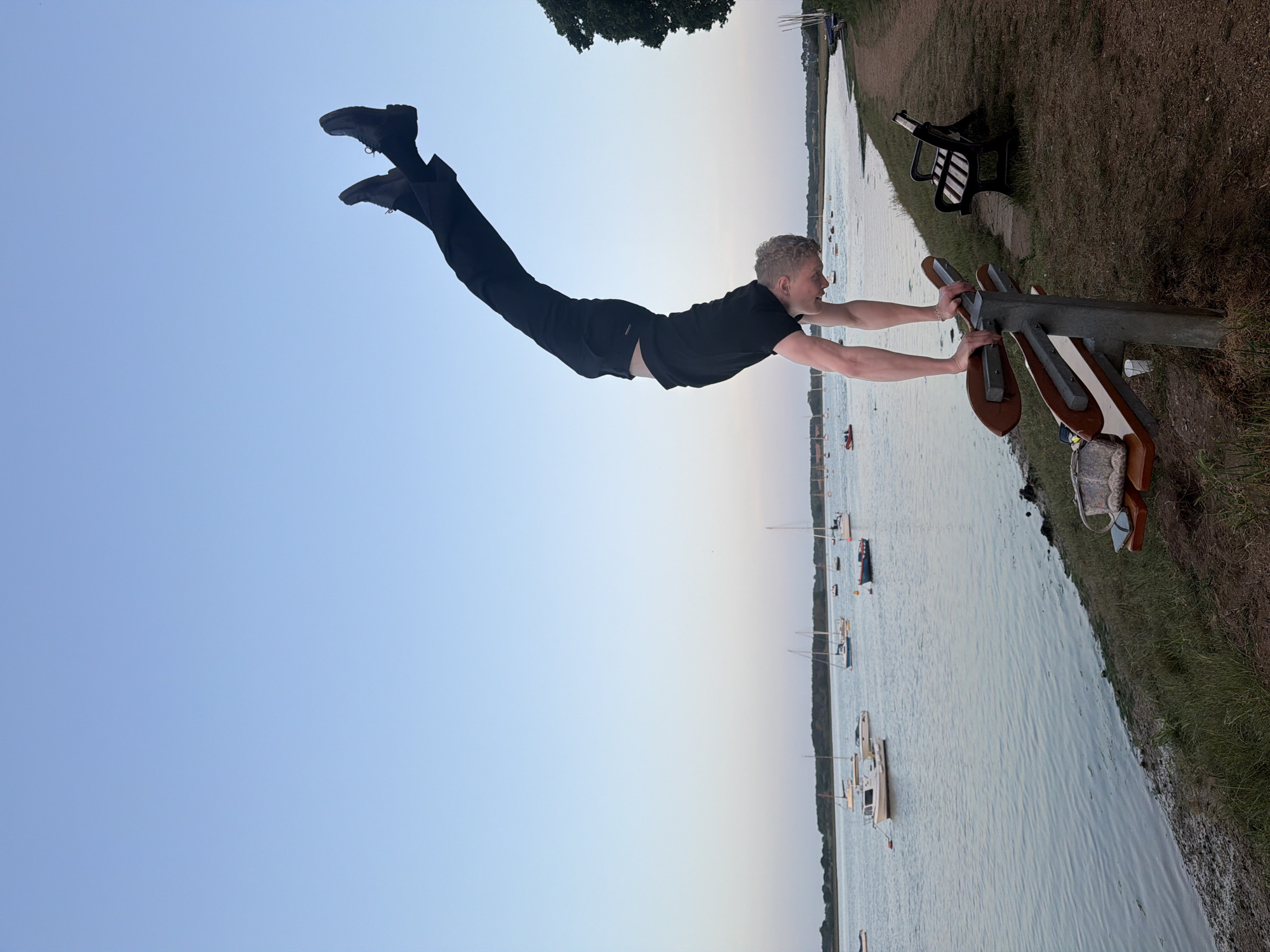 A man is photographed parallel to the ground, holding onto a metal sculpture in an outdoor setting. He is wearing black trousers, a black t-shirt, and black shoes. He has short blonde hair. The sculpture appears to be a stylized depiction of an airplane. The background features a body of water with several boats moored in it. Beyond the water is land with vegetation. A bench sits on the grassy bank. The sky is a pale blue. The location is in East Suffolk, United Kingdom.
