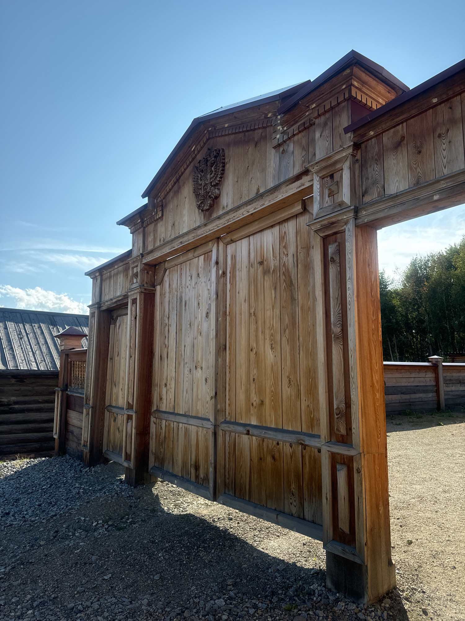 A low-angle outdoor shot captures a large, rustic wooden gate, likely an entrance to a historical park or settlement, in the city of Taltsy (Irkutsk), Russia. The gate, constructed from vertical planks of aged wood, features intricate carvings and decorative elements, including what appears to be a double-headed eagle emblem prominently displayed above the central opening. The gate is flanked by substantial wooden pillars, and a smaller doorway is visible within the left-hand side of the main gate. The ground in front of the gate is covered in gravel, with shadows cast by the structure indicating a sunny day. Beyond the gate, a wooden fence and trees are visible, suggesting a rural or park-like setting. The sky above is a clear, bright blue with a few wispy clouds. The overall impression is one of historical architecture and an outdoor, possibly tourist-oriented, location.