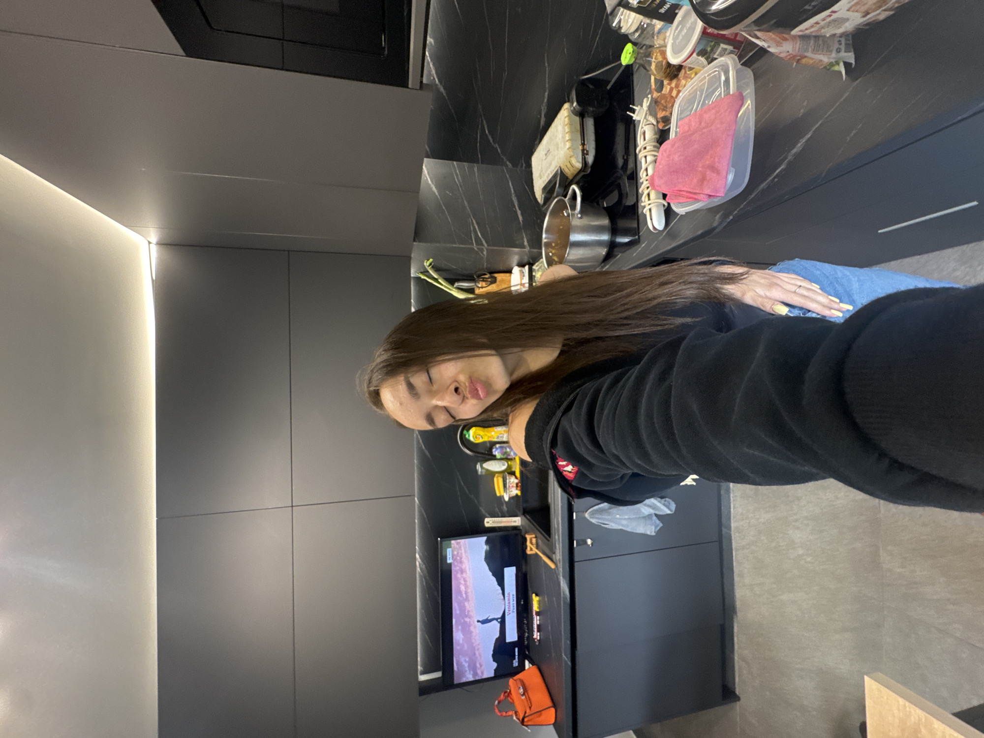 The image captures a young woman taking a self-portrait in a modern kitchen, likely in Sofia, Bulgaria. The woman, with long brown hair, is wearing a black hoodie and blue jeans. Her left hand, adorned with yellow nail polish and a ring, rests casually on her leg. She gazes towards the camera with a relaxed, slightly pensive expression, her eyes appearing partially closed.
The kitchen features a sleek, contemporary design dominated by dark elements. The cabinetry is a matte dark gray or black, seamlessly integrated into the wall. The countertop is a dark, possibly marble or granite, surface with subtle lighter veining. Various kitchen items are scattered across it, including a metal pot, plastic containers (one holding a pink cloth), and other cooking utensils, suggesting a lived-in space.
A small television screen sits on the countertop to the right of the woman, displaying a scene with a person silhouetted against a landscape, and the word "Venomous" along with another partly visible word like "kurt" (possibly part of a name or title) underneath. An orange, possibly leather, bag is placed on the counter beneath the TV. The floor appears to be covered with large, light-colored tiles. Integrated strip lighting runs along the top of the wall cabinets and possibly under them, casting a warm glow and highlighting the clean lines of the kitchen architecture. The overall ambiance is modern and urban, illuminated by artificial lighting, suggesting an indoor setting at any time of day.